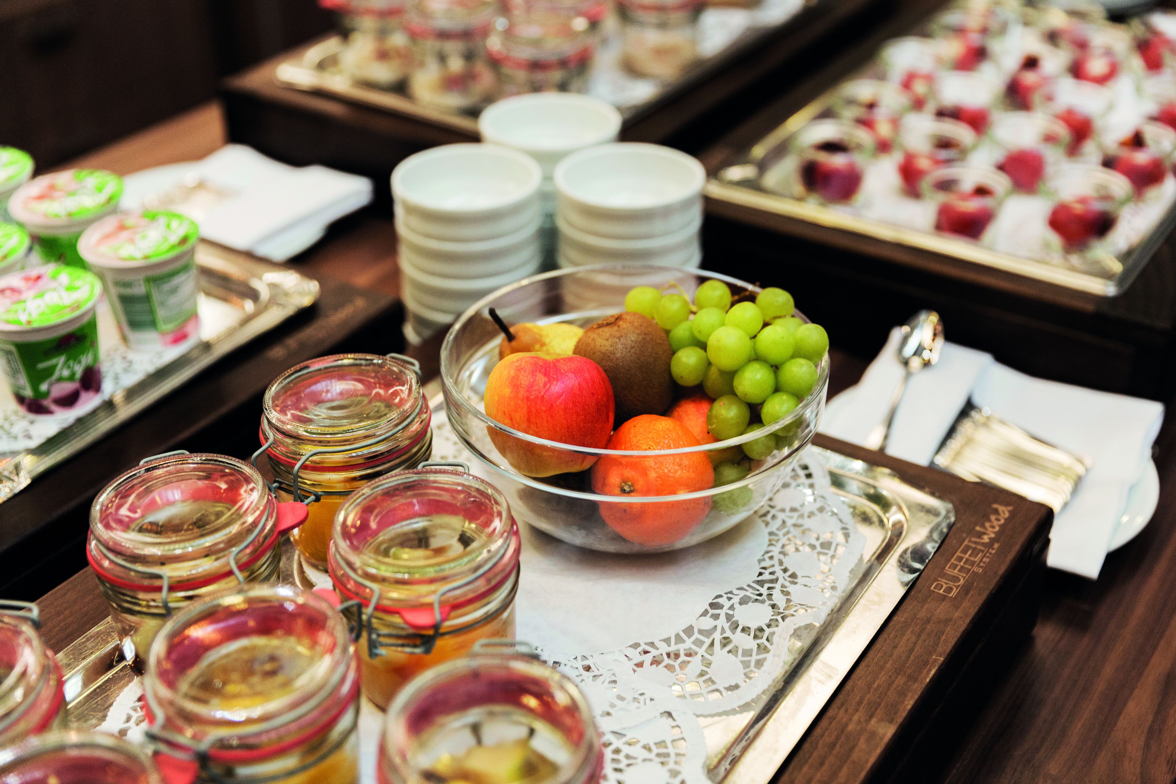 Uma mesa de buffet com diversos alimentos, incluindo uma tigela de frutas, potes de compotas e pratos empilhados em um suporte.