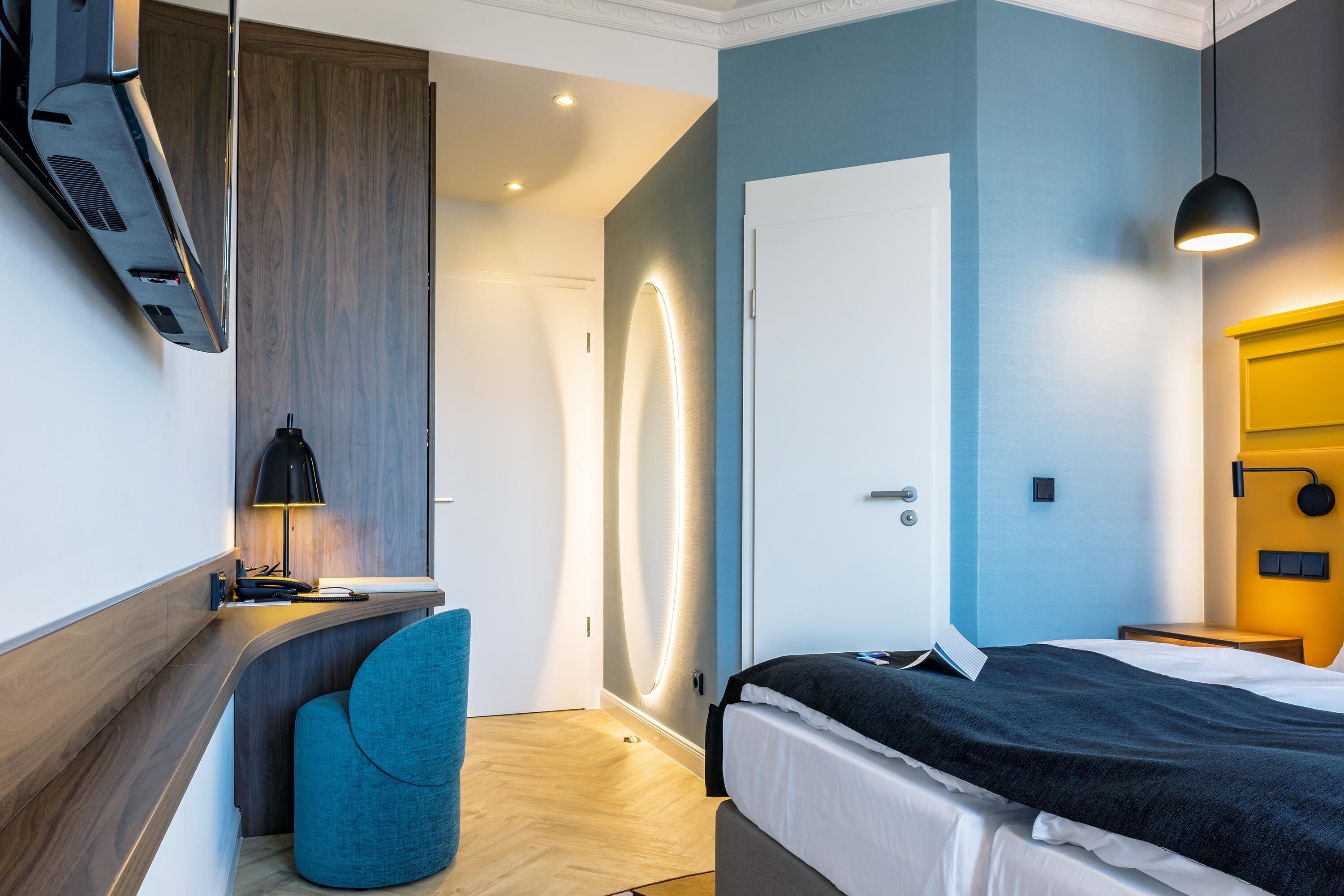 Een moderne hotelkamer met een gebogen bureau, een comfortabele fauteuil, een bed met een zwarte plaid en een heldere wandlamp.