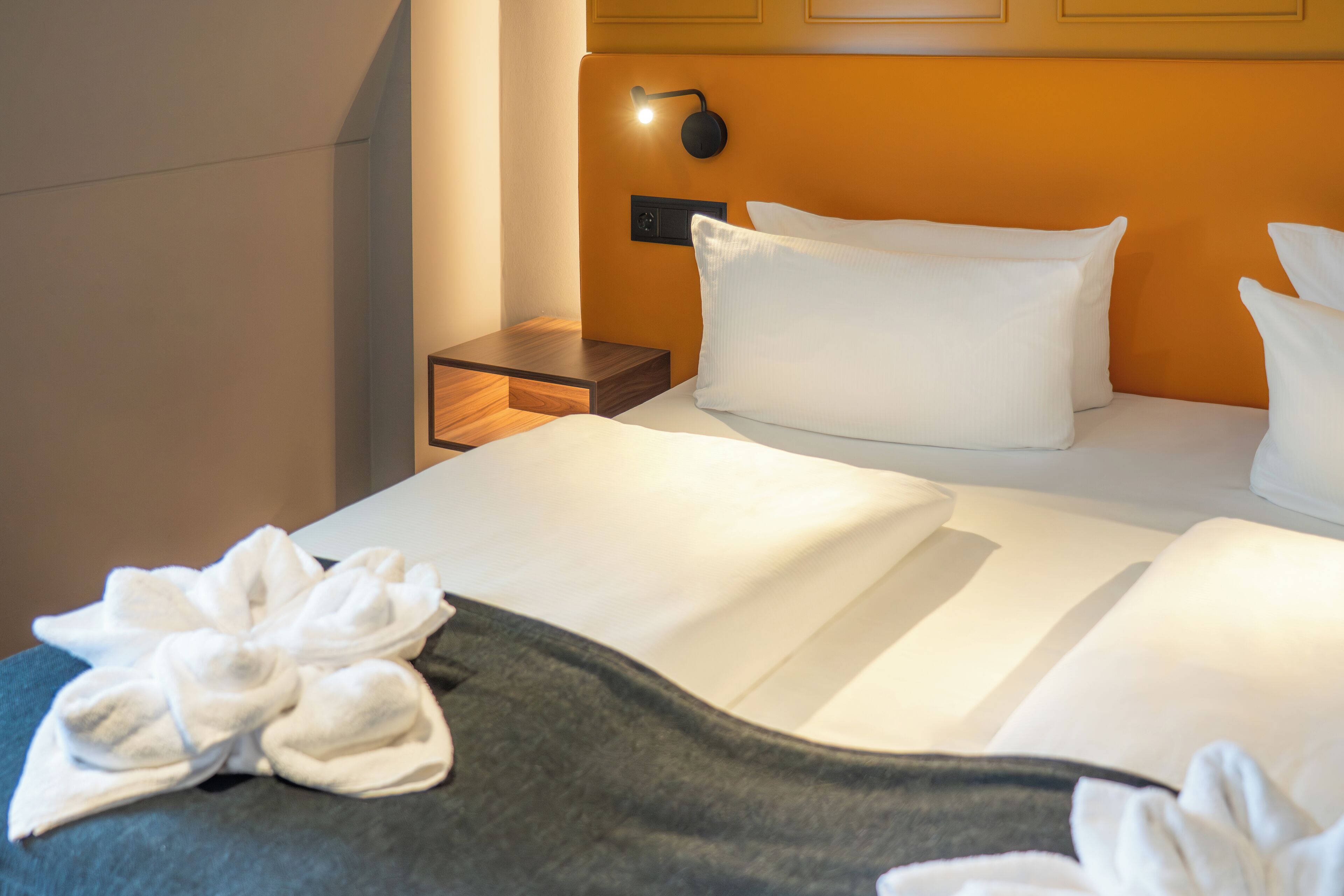 Een net opgemaakt bed met witte lakens en een donkere onderrok, een nachtkastje met een lamp, en een wandlamp.