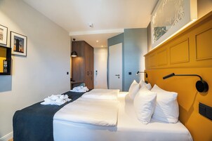 Habitación de hotel con dos camas, un cabecero oscuro, una lámpara de pared y una puerta que da a otra habitación.