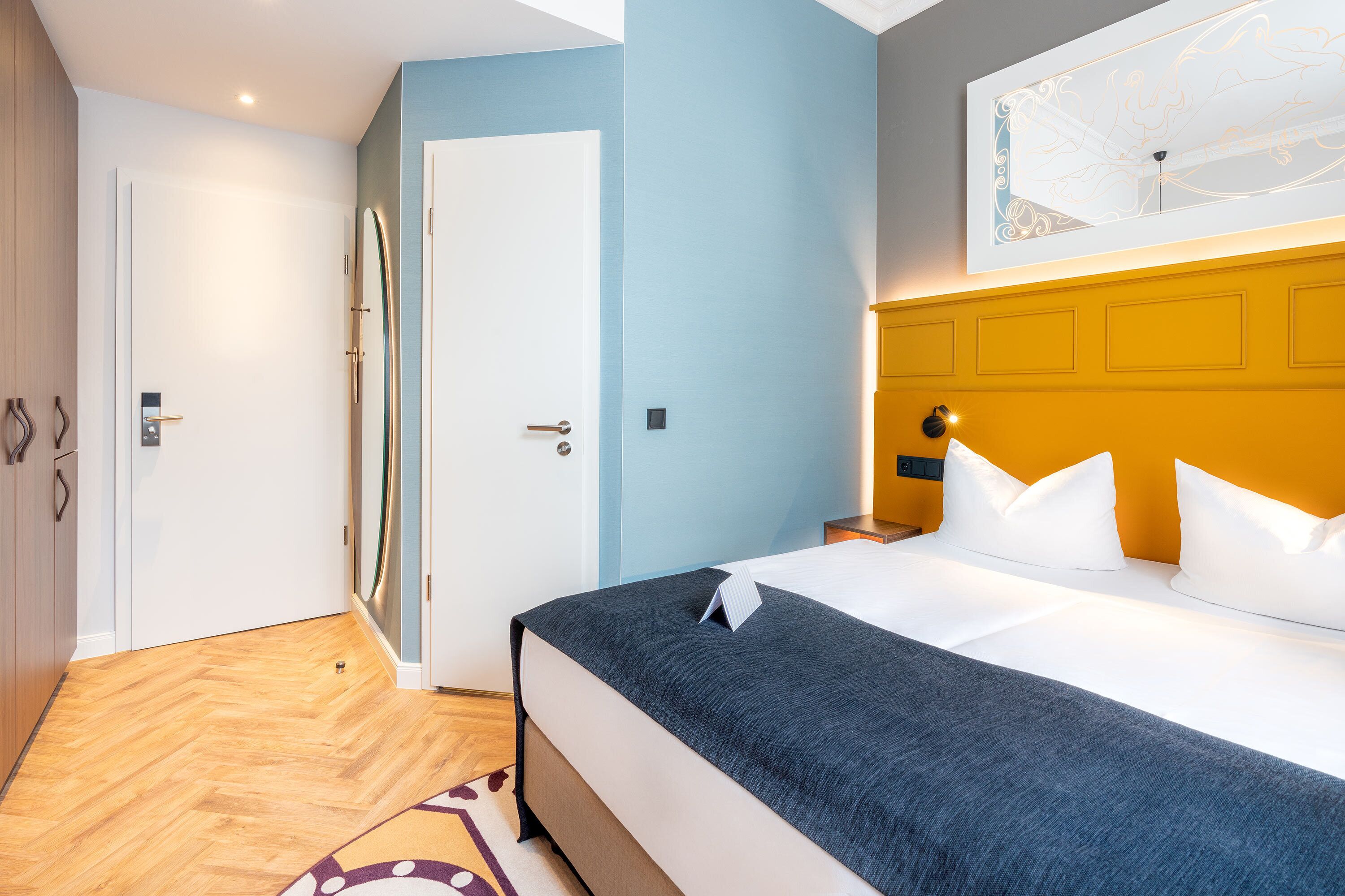 A hotel room with a bed, a nightstand, a door, and a wall with a light fixture.