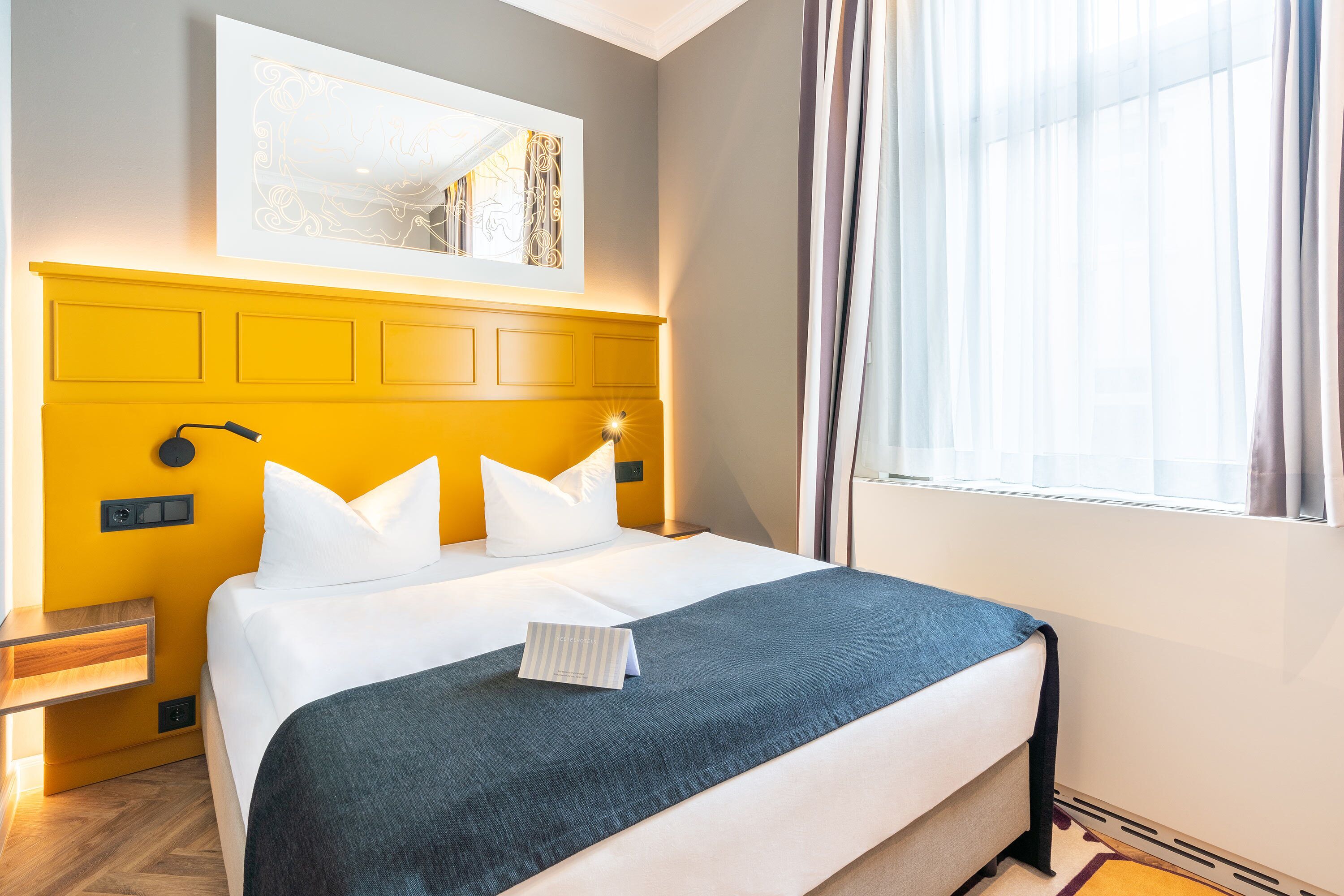 Habitación de hotel con cama, mesita de noche, lámpara de pared y ventana con cortinas.