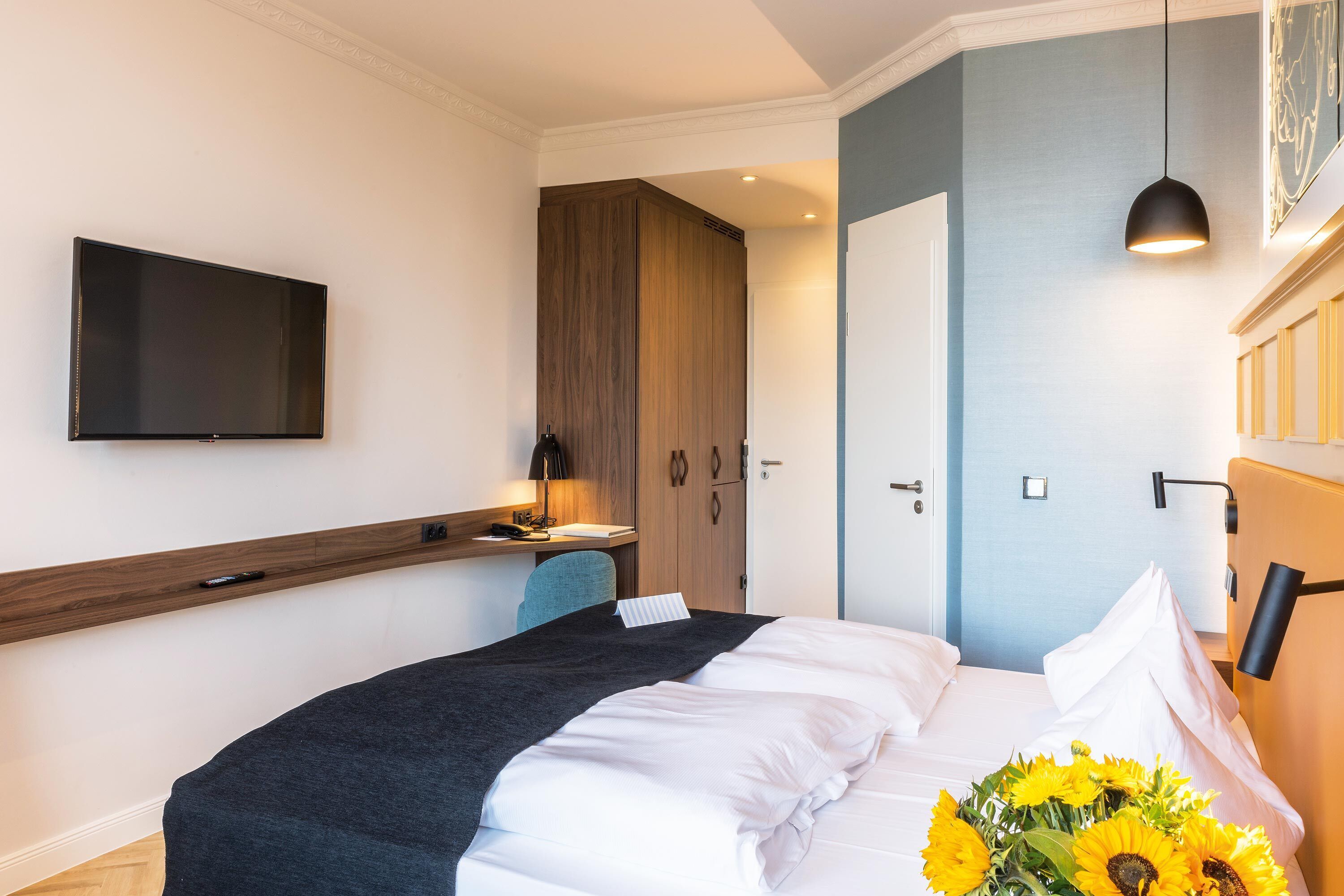 Una habitación de hotel moderna con una cama grande, un televisor de pantalla plana en la pared, un escritorio con lámpara y una puerta que da a otra habitación.