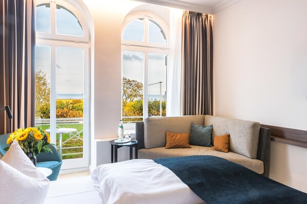 Une chambre d’hôtel avec un lit, un canapé, une petite table sur laquelle est posé un vase de fleurs, et de grandes fenêtres donnant sur l’extérieur.