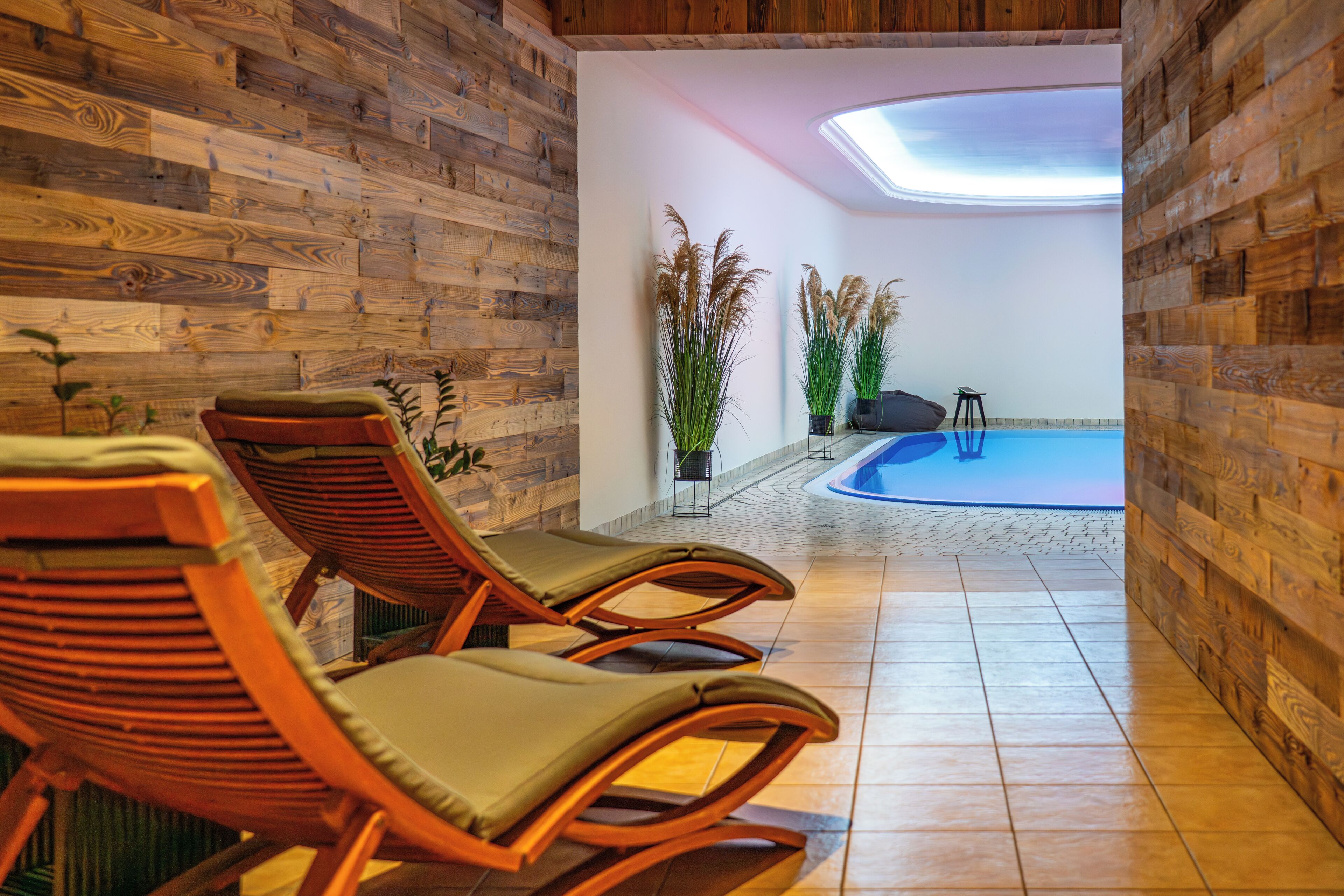 A room with wooden walls, a tiled floor, and a pool area. There are lounge chairs and potted plants.