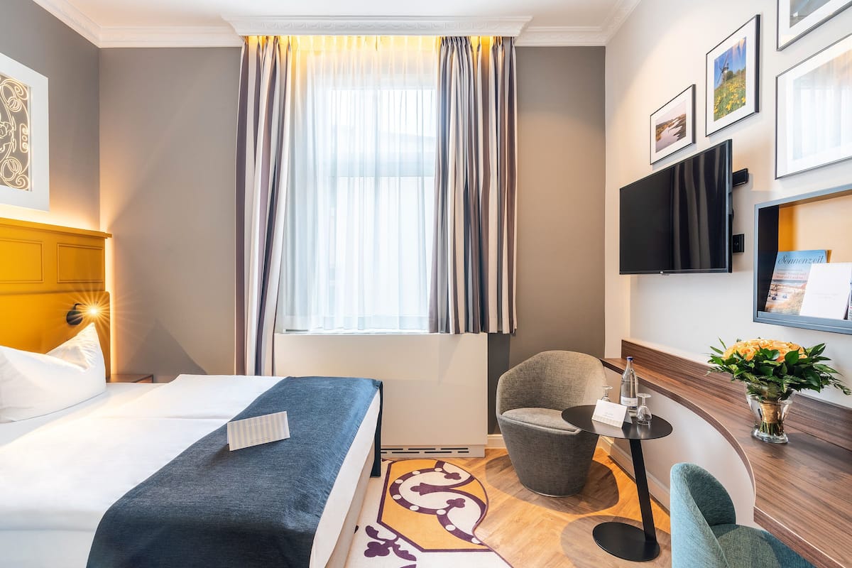 A hotel room with a bed, a chair, a small table with a vase of flowers, and a flat-screen TV mounted on the wall.