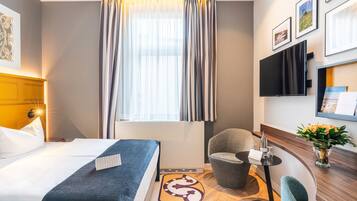 A hotel room with a bed, a chair, a small table with a vase of flowers, and a flat-screen TV mounted on the wall.