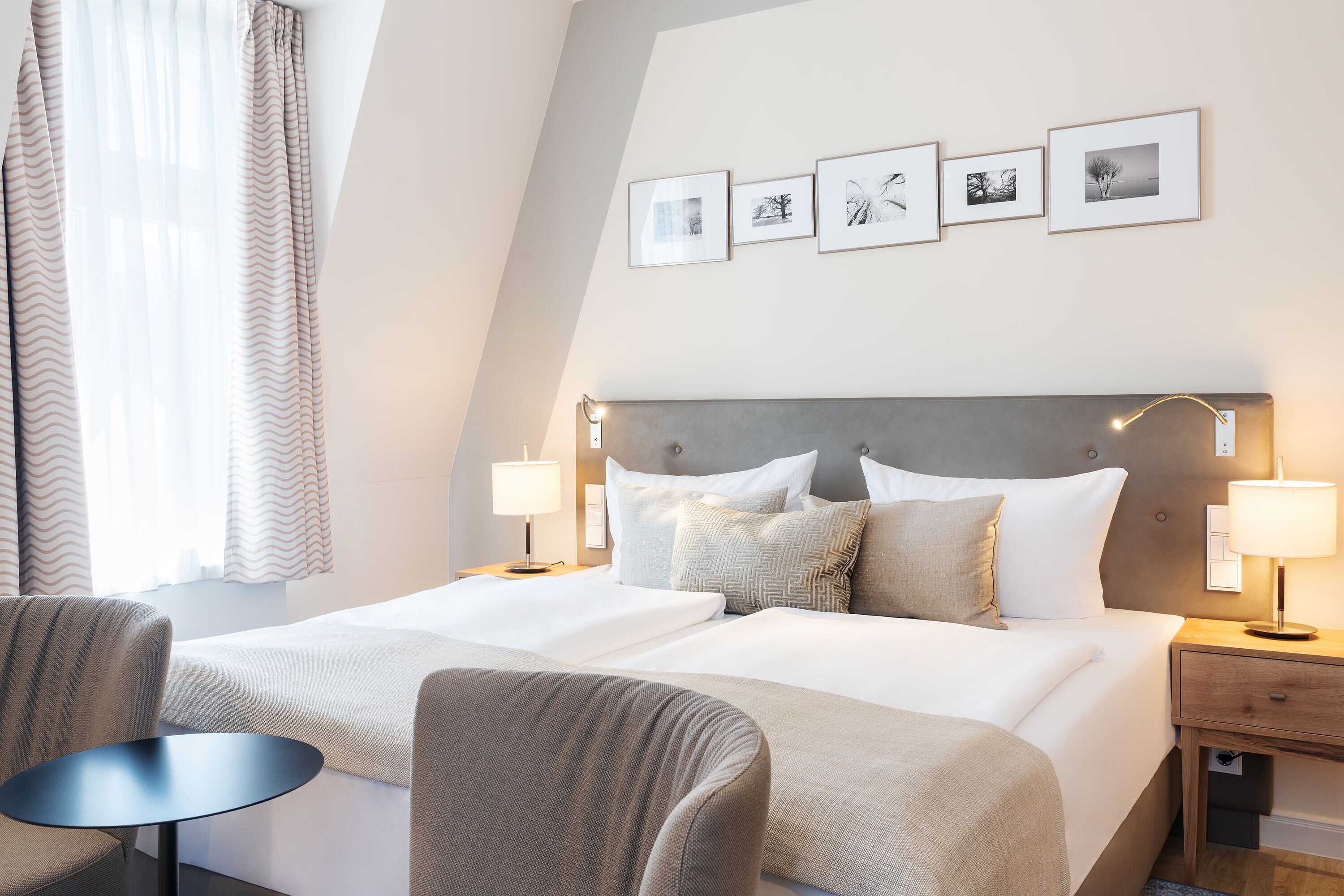 Ein Hotelzimmer mit einem großen Bett, zwei Nachttischen, einem Sessel und einem kleinen Tisch. An der Wand hängen vier gerahmte Bilder.