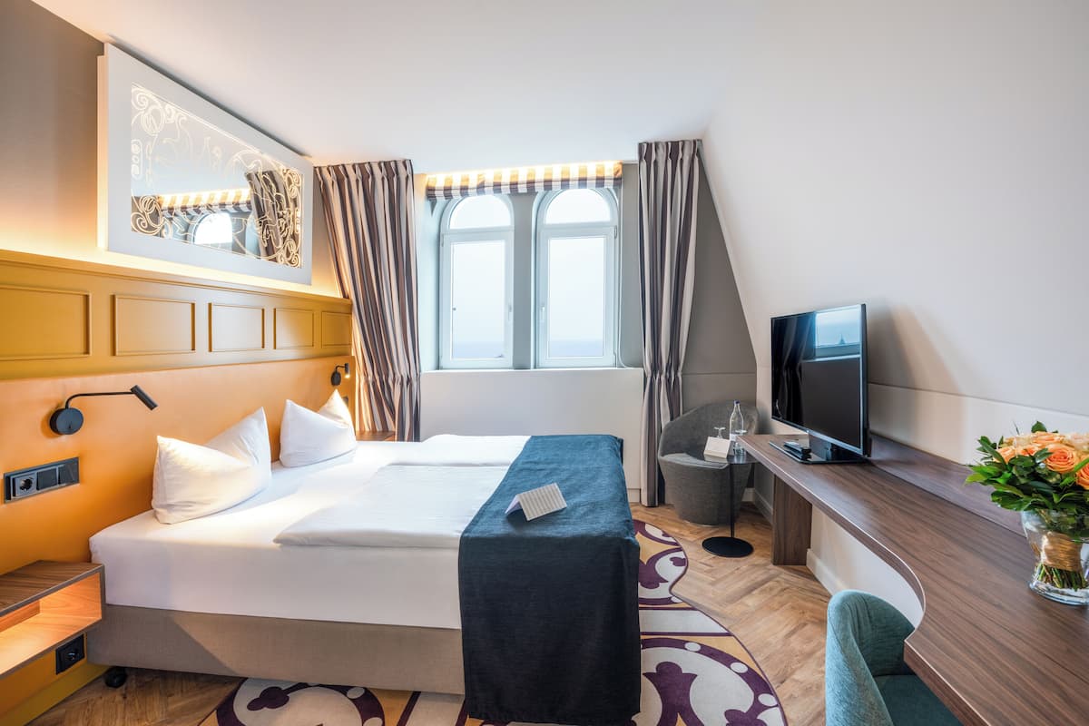A hotel room with a bed, a desk with a TV, a window with curtains, and a wall-mounted light fixture.