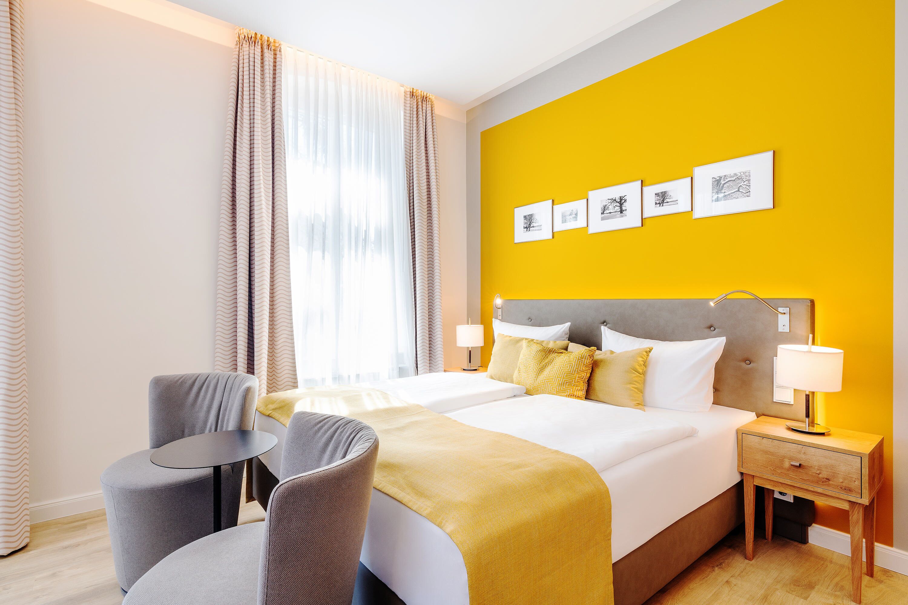 Una habitación de hotel con cama, dos mesitas de noche, una silla, ventana con cortinas y una obra de arte en la pared.
