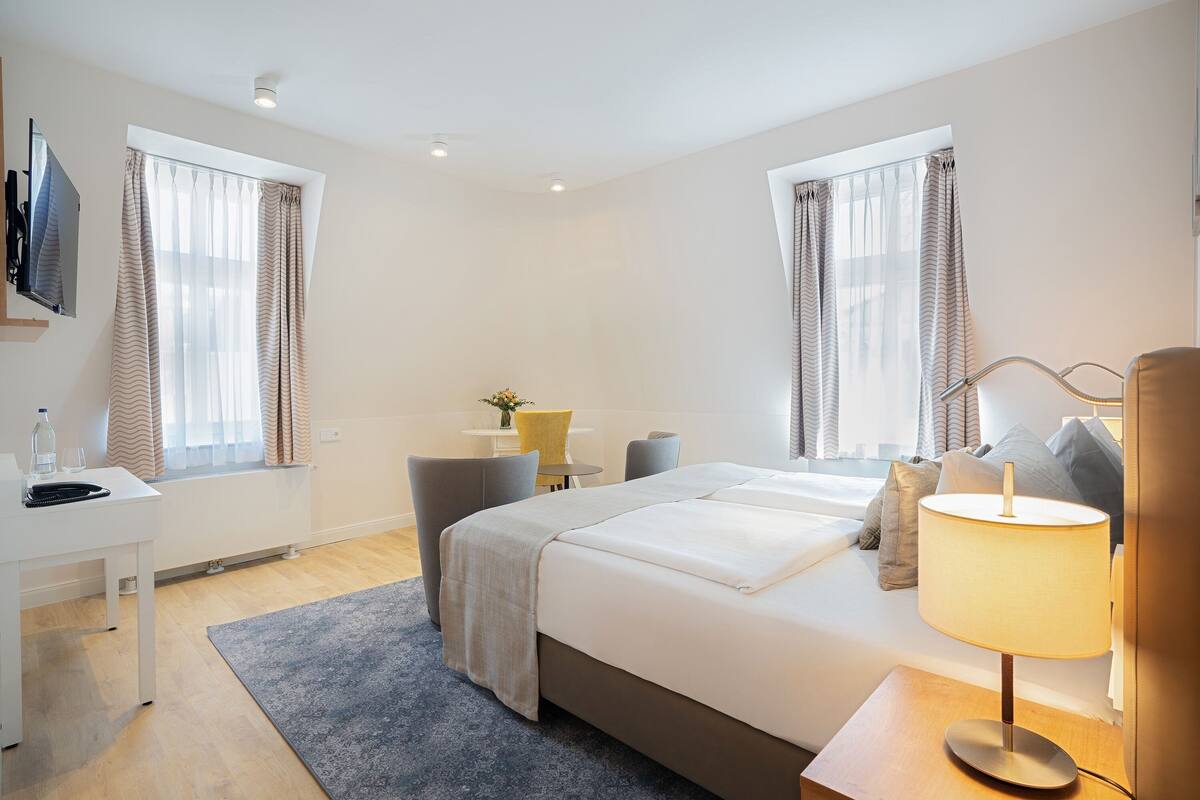 A modern hotel room with a large bed, a desk, a chair, a lamp, and a small table with a vase of flowers.