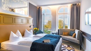 A hotel room with a large bed, a sofa, a small table, and a view of the outdoors through large windows. - SEETELHOTEL Ostseehotel Ahlbeck (Heringsdorf)