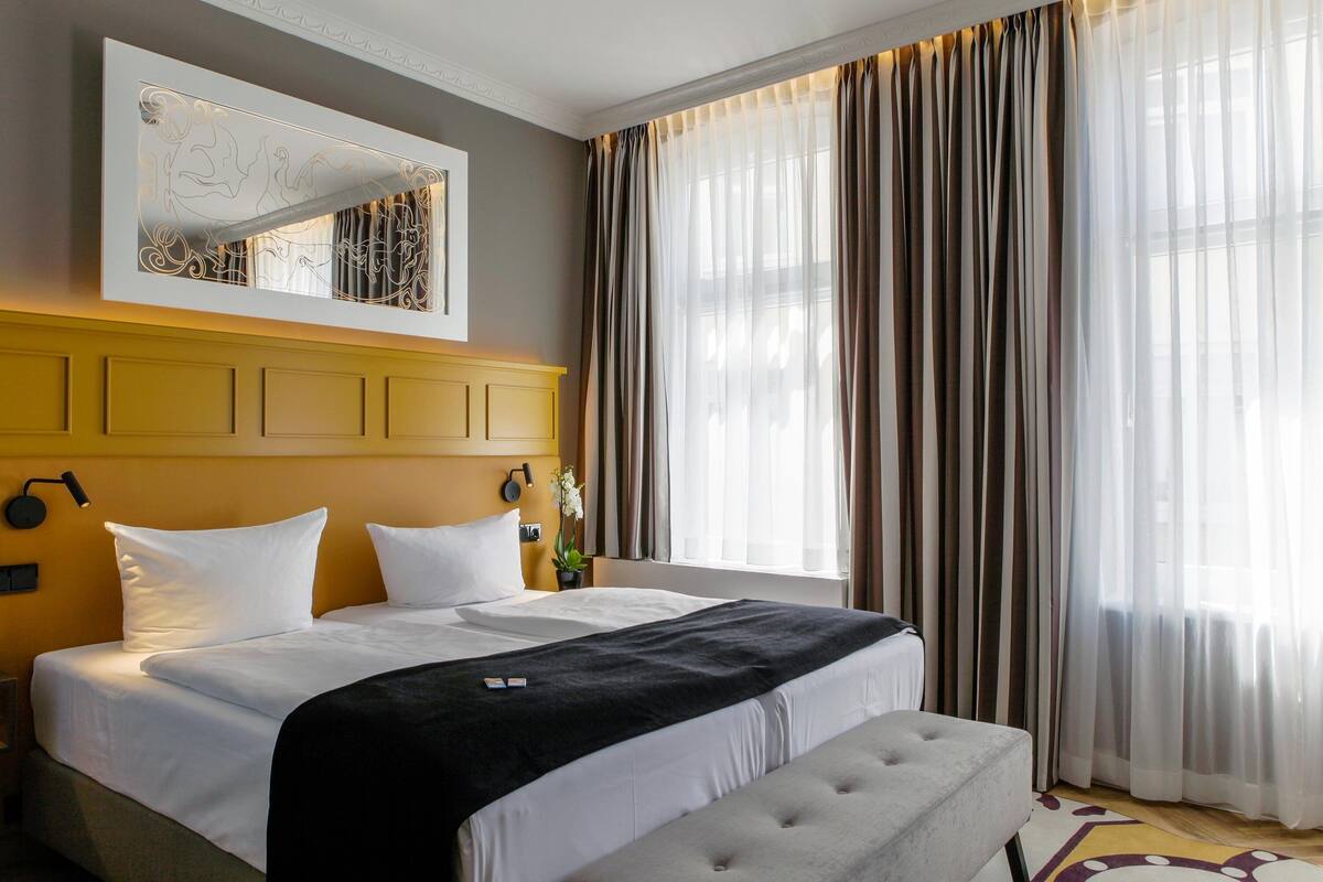 A hotel room with a large bed, a headboard with a mirror, a bedside table with a lamp, and a window with curtains.