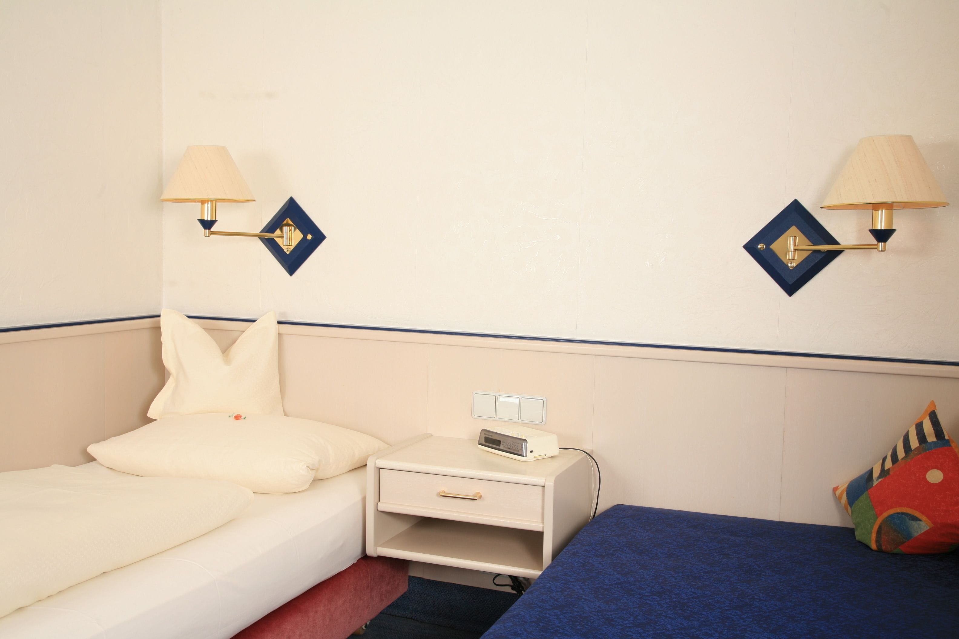 A hotel room with a bed, bedside table, lamp, and a small drawer unit.