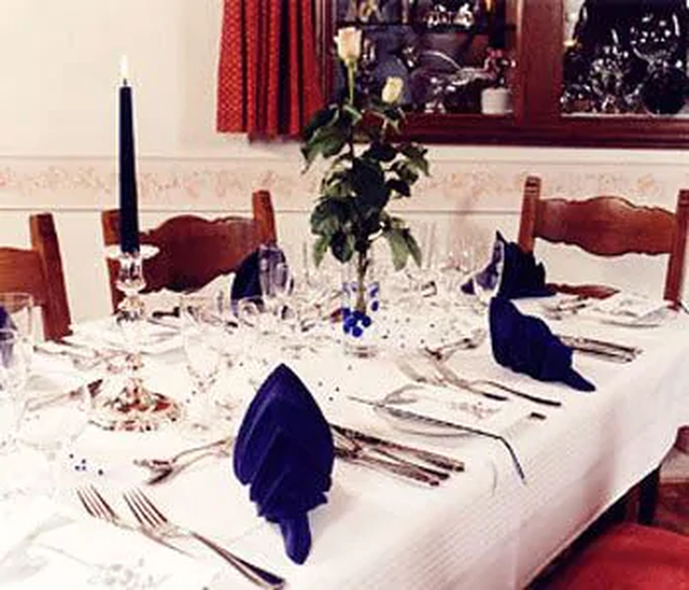 Ein Esstisch ist für eine Mahlzeit gedeckt, mit weißer Tischdecke, blauen Servietten und Besteck.