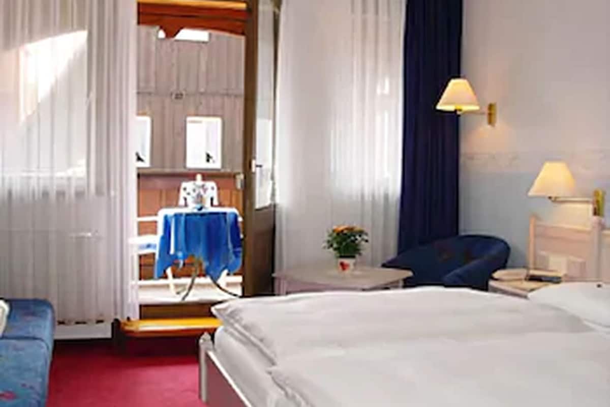 Habitación de hotel con una cama, una silla, una mesita con lámpara y vistas a una zona de cocina.