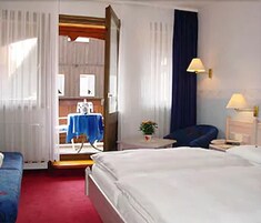 Quarto de hotel com uma cama, uma cadeira, uma mesa pequena com abajur e vista para uma cozinha.