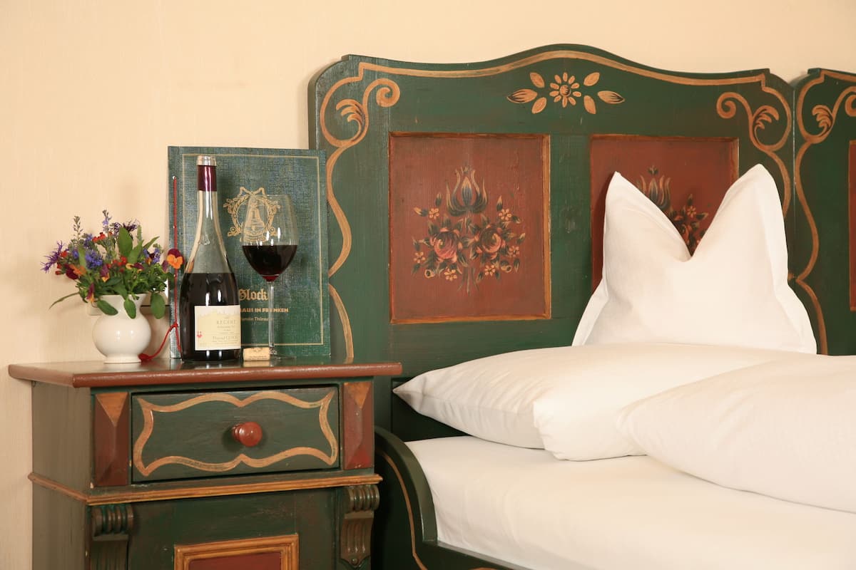Una cama bien hecha con sábanas blancas, una mesita de noche de madera con una botella de vino y un florero, y un cabecero decorativo.