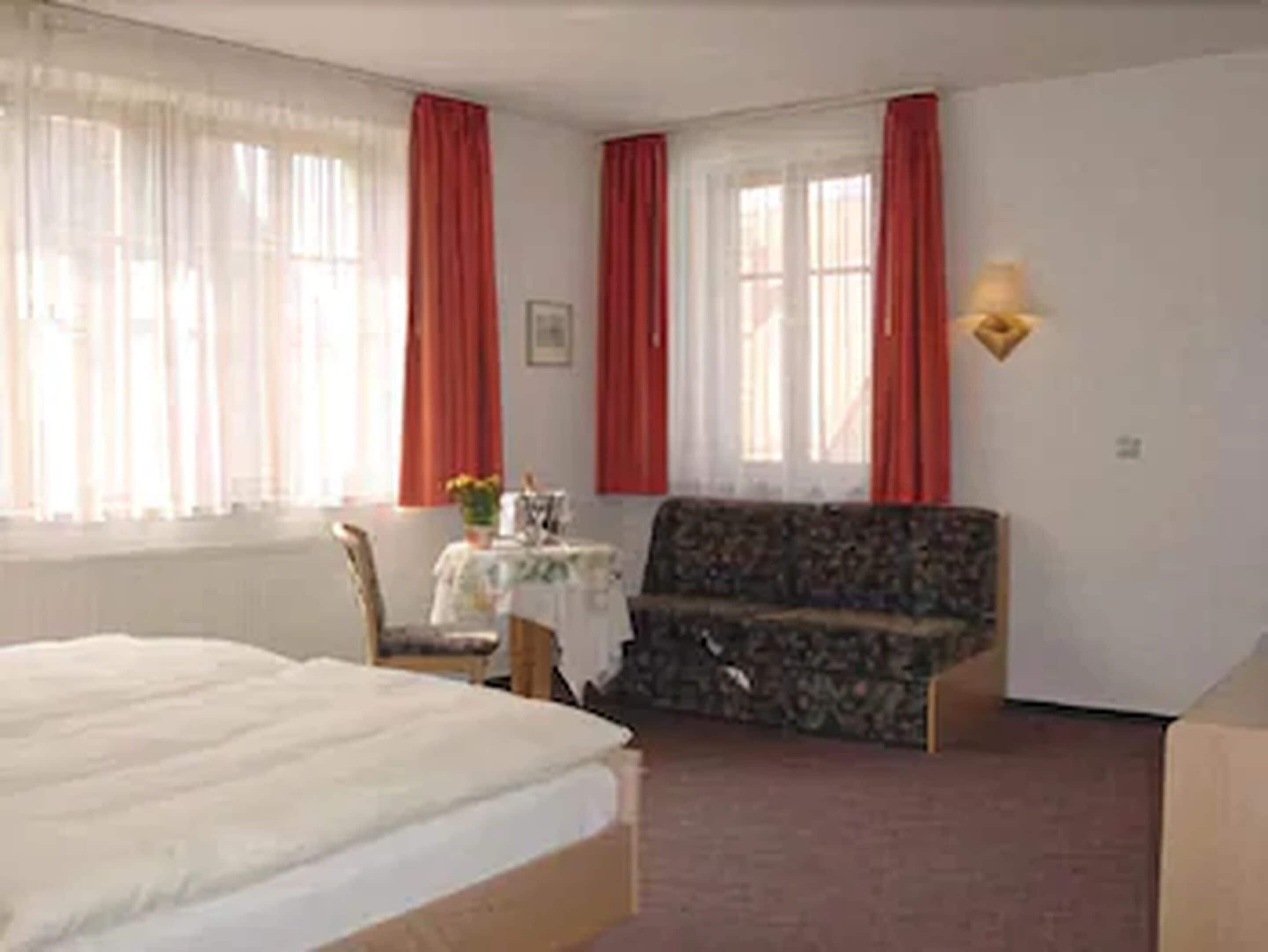 Ein Schlafzimmer mit Bett, Sofa, Sessel, einem kleinen Tisch mit Vase und einem Fenster mit Vorhängen.