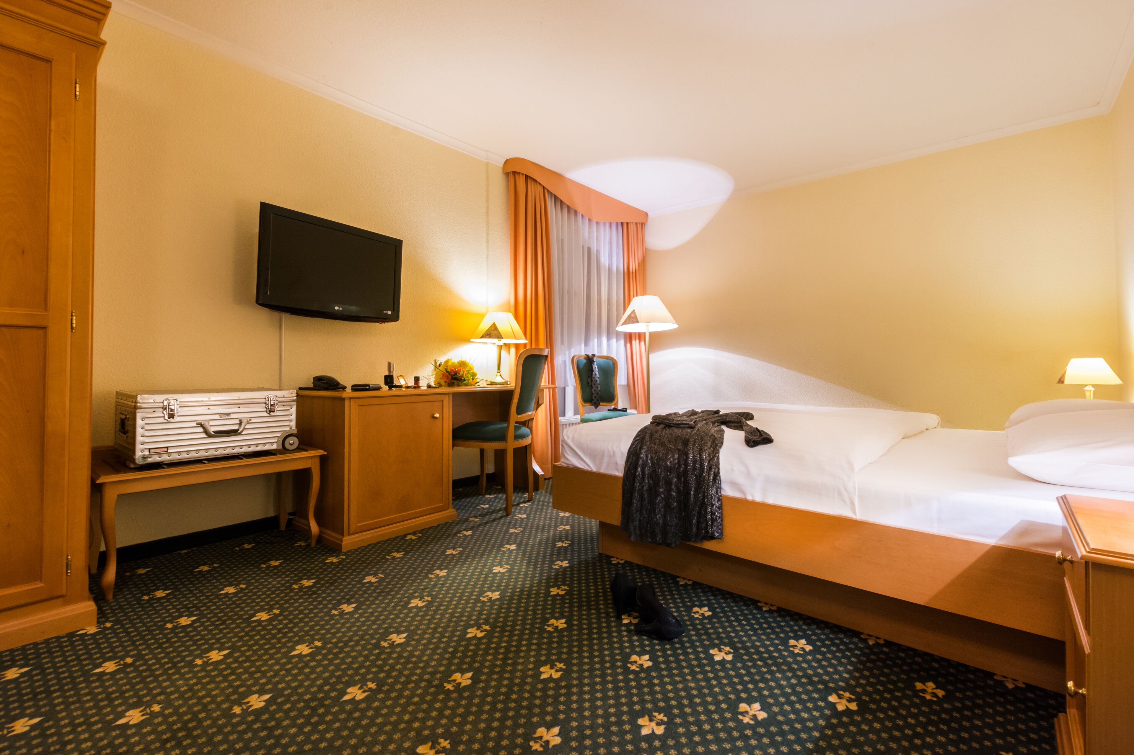 A hotel room with a bed, a desk with a suitcase, a television, and two lamps.