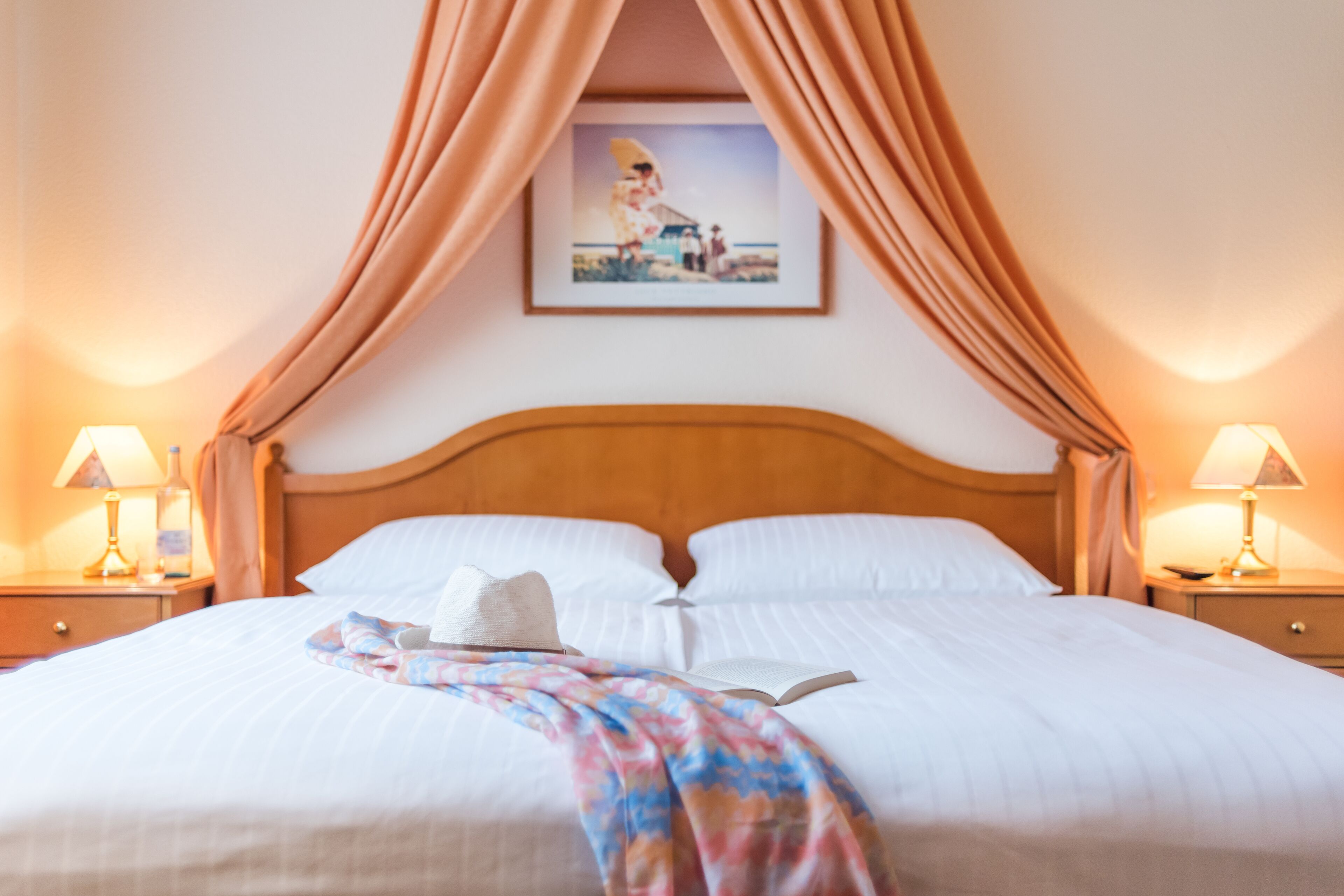 A neatly made bed with white linens, a floral throw blanket, and a hat on the pillows. There are two bedside tables with lamps and a framed picture above the bed.