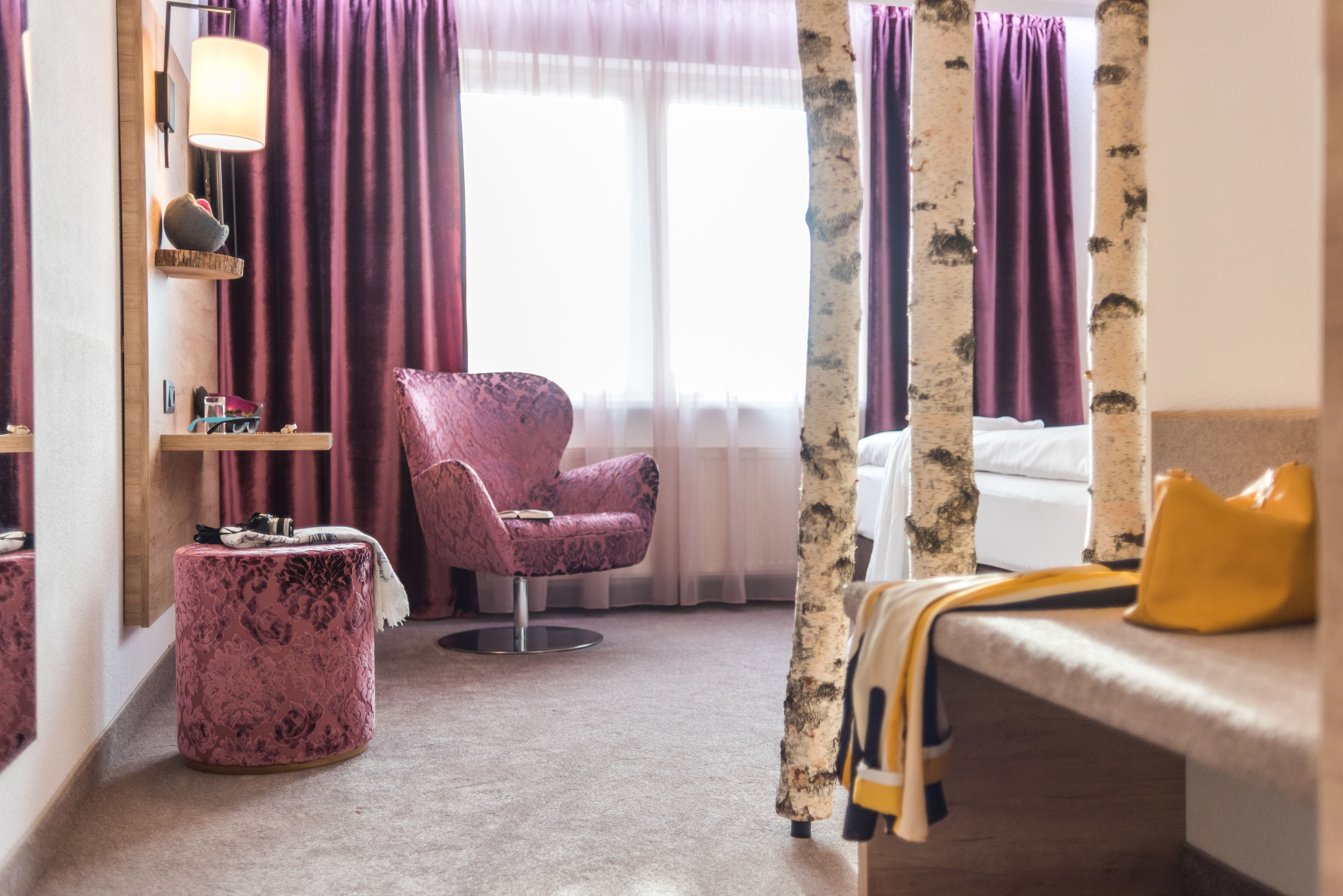Una habitación de hotel con una cama, un sillón rosa, un otoman redondo, un espejo y una ventana con cortinas.