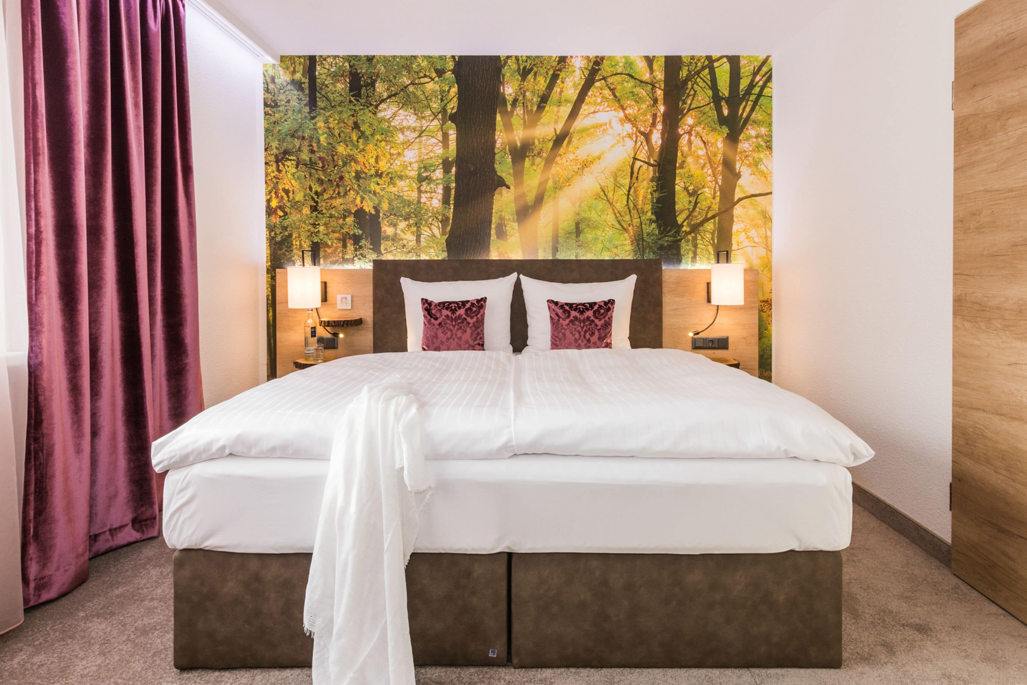 Ein Hotelzimmer mit einem grossen Bett, zwei Nachttischlampen, einem Kopfteil mit einem Waldgemälde und einer Holztür.