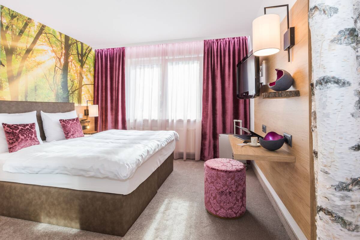 Ein Hotelzimmer mit einem großen Bett, einem Holztisch, einem Spiegel, einem runden pinkfarbenen Pouf und einem an der Wand befestigten Fernseher.