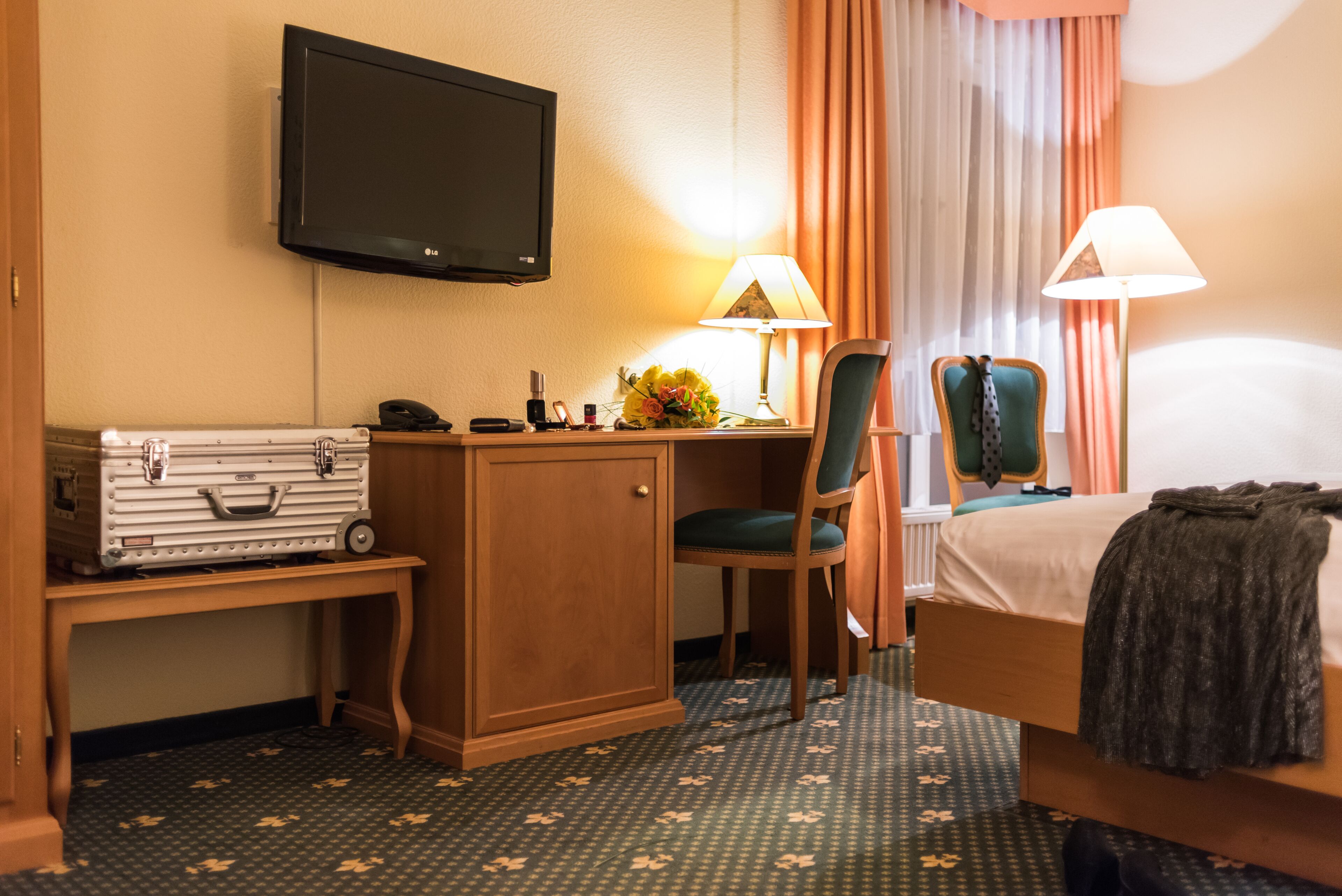 A hotel room with a bed, desk, chair, television, and a small table with a lamp and flowers.