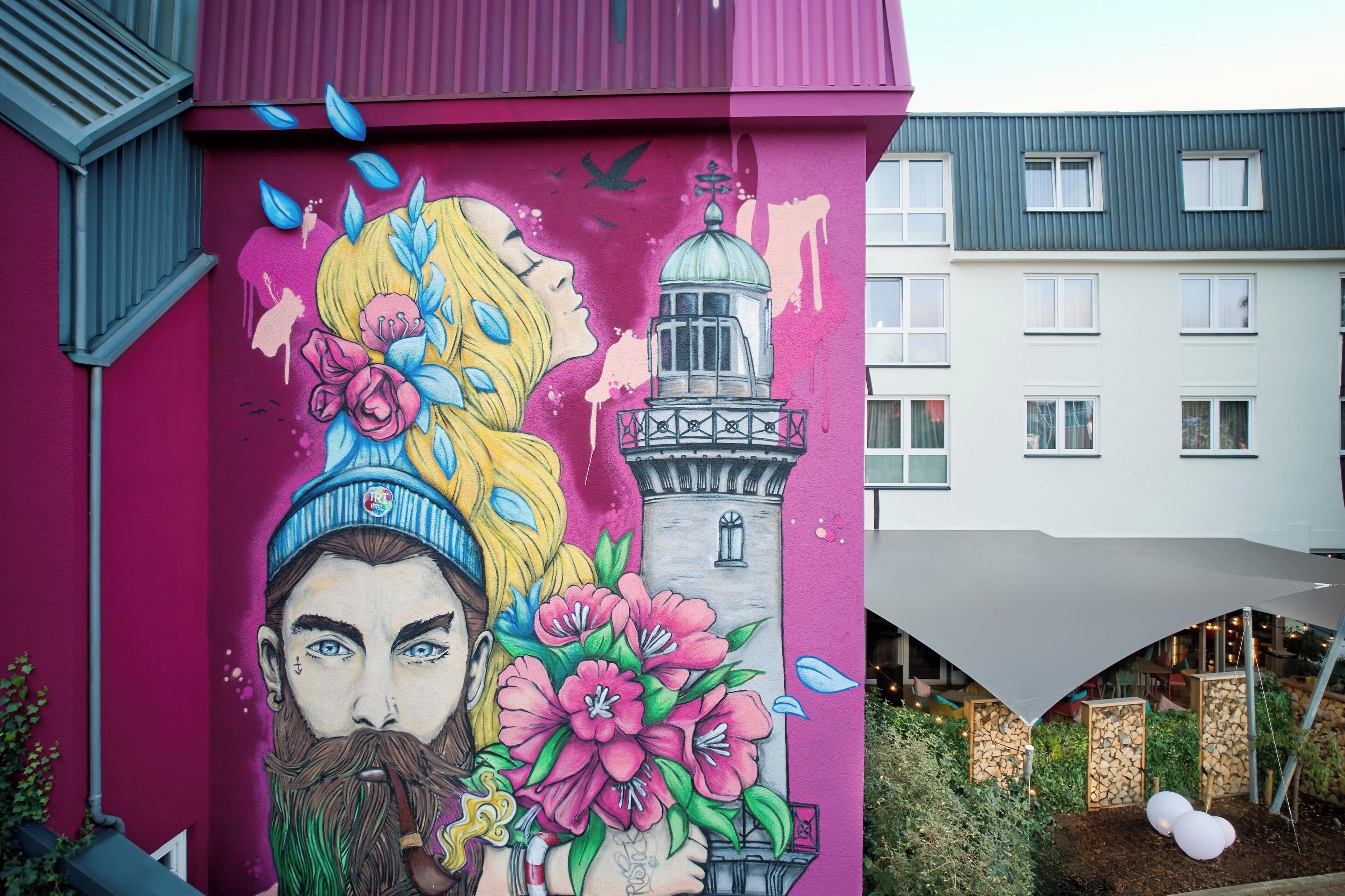 Un mural colorido en un edificio con una torre, un toldo rosa y un jardín con globos.