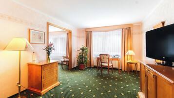Habitación de hotel con alfombra floral verde, cómoda de madera, televisor, escritorio con silla y un espejo grande.