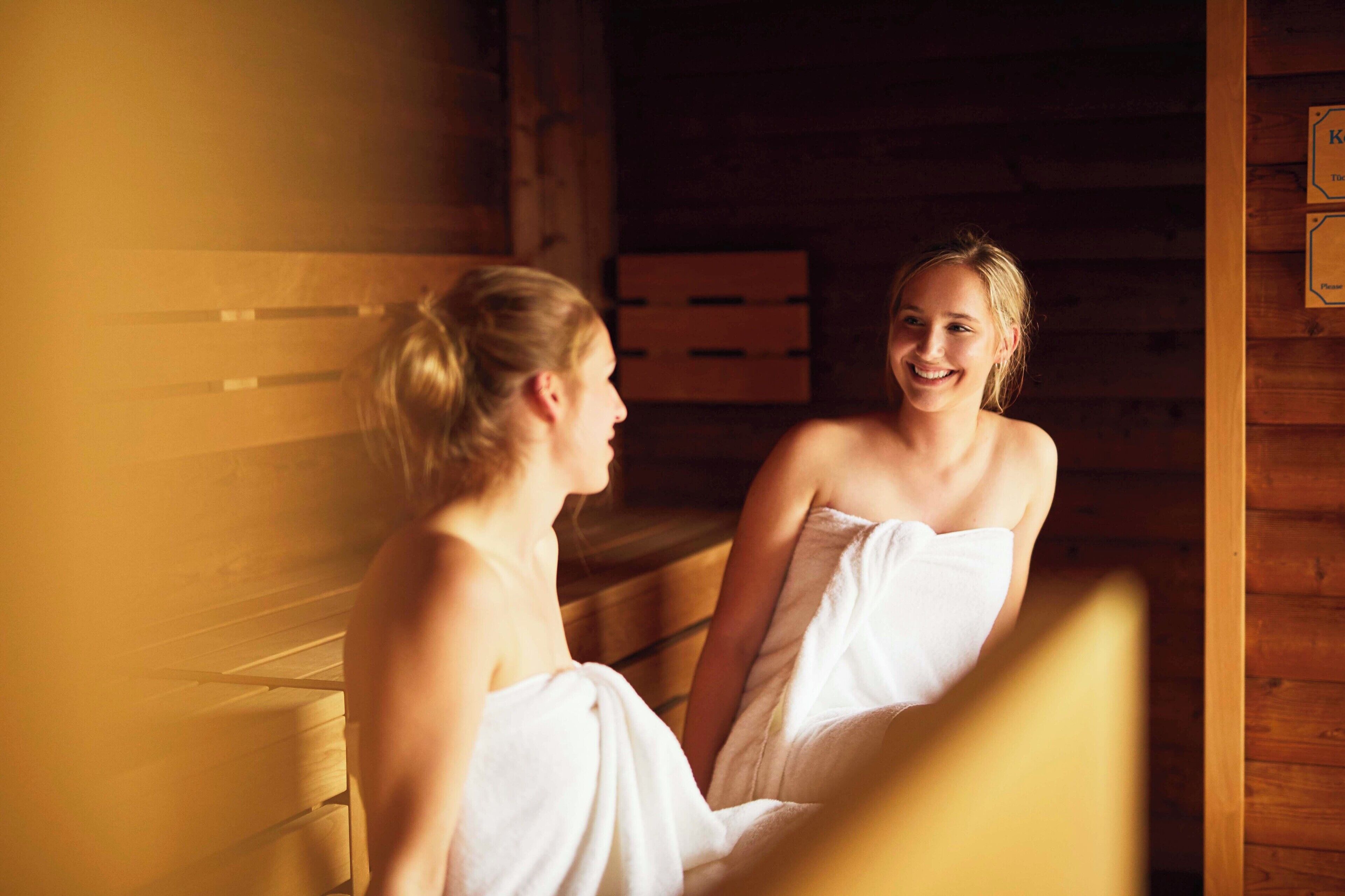 Dos personas sentadas en una sauna de madera con bancos, envueltas en toallas.