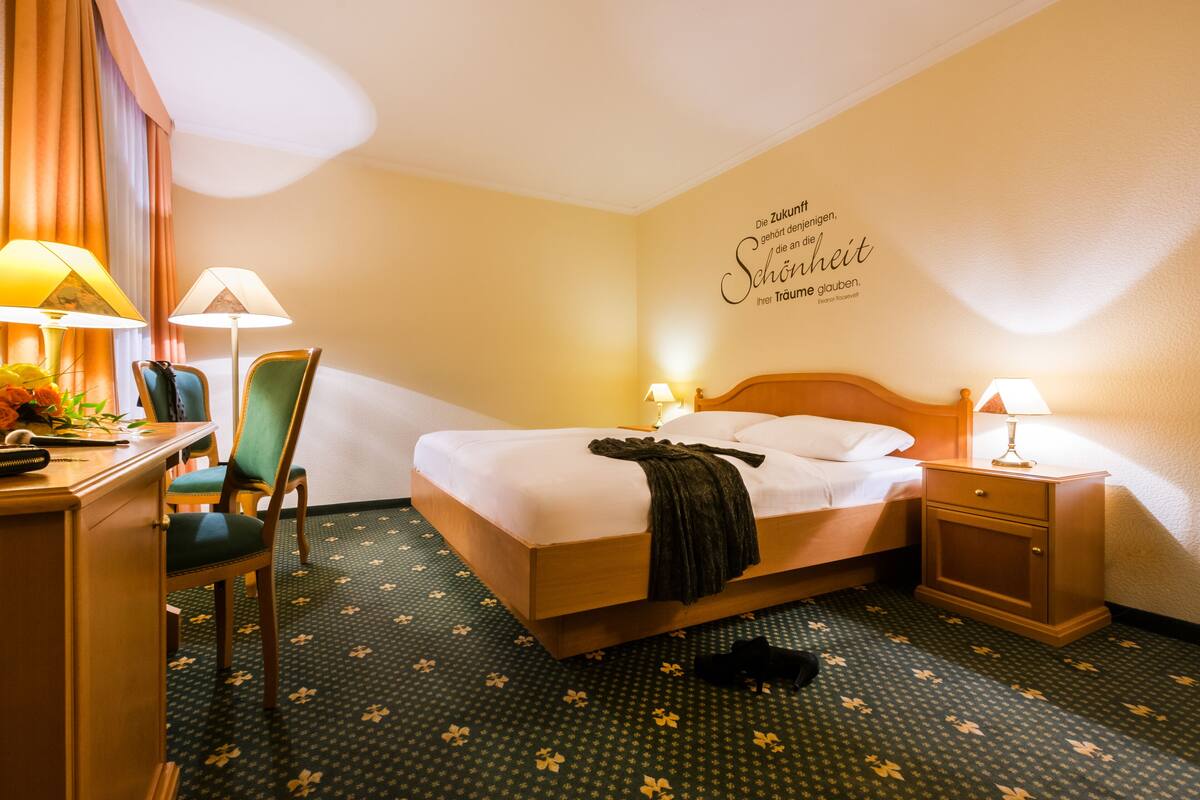 Ein Hotelzimmer mit einem Bett, zwei Nachttischen mit Lampen, einem Schreibtisch mit Stuhl und einer Wand mit einer dekorativen Schrift.