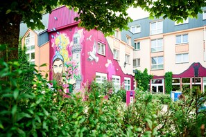 Vibrant building with a colorful mural and lush greenery surrounds a residential space with multiple windows and balconies.
