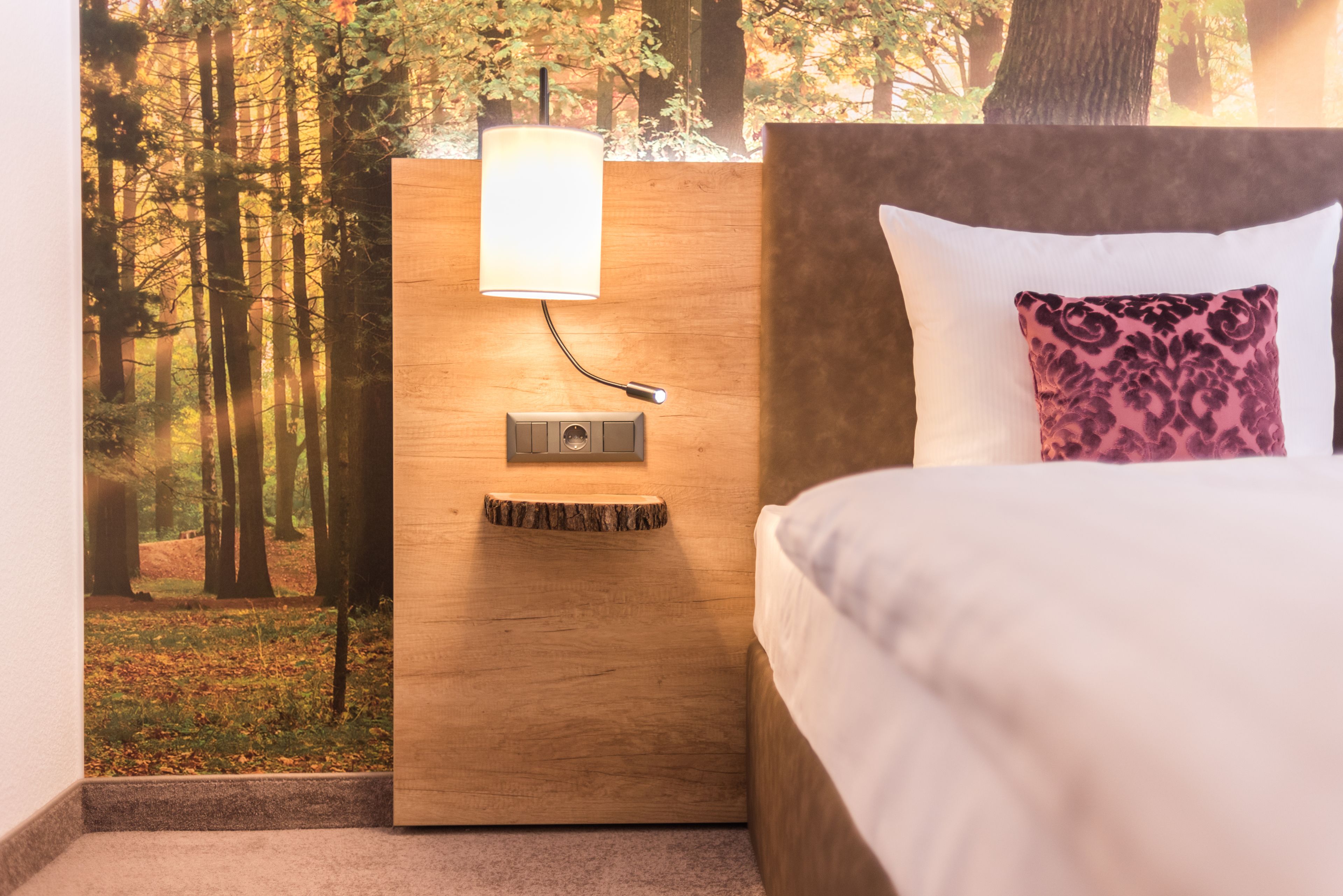 A bedroom with a bed, a nightstand with a lamp, and a wall mural of a forest.