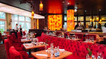 Un restaurant avec des banquettes en velours rouge et des tables en bois, un coin bar et une grande fenêtre laissant entrer la lumière naturelle.