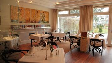 A dining area with tables set for service, a large window with curtains, and a wall with patterned artwork.