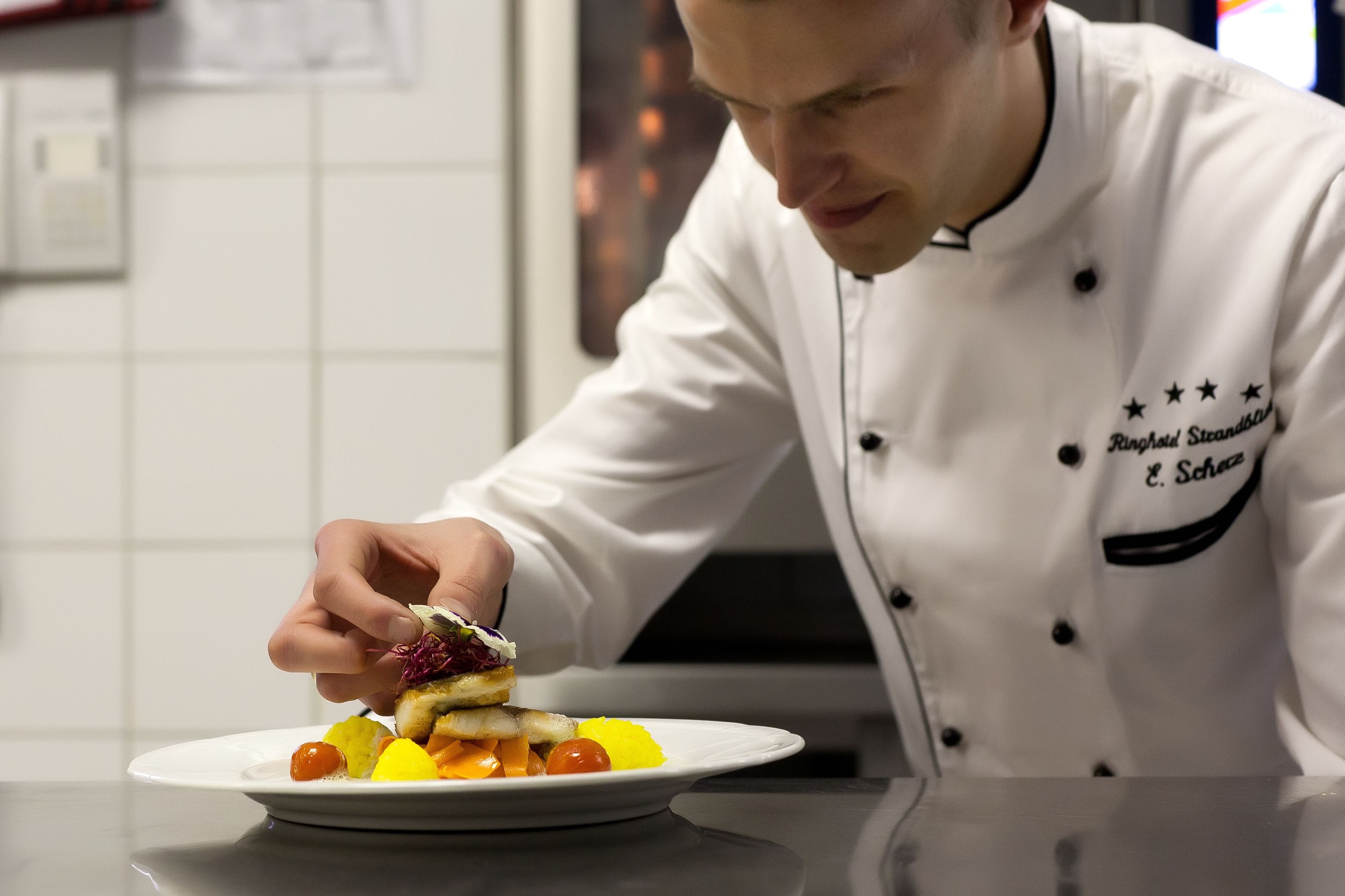 A chef in a white uniform with a name tag, preparing a dish on a white plate with various ingredients.