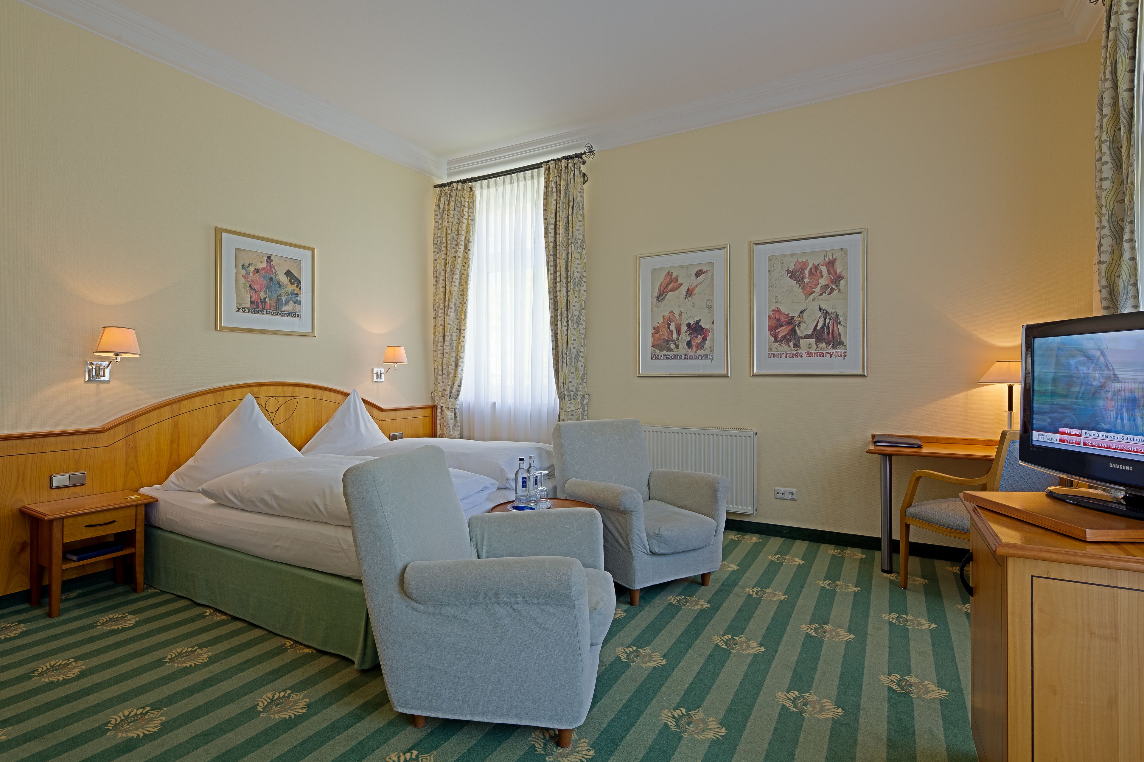 Ein Hotelzimmer mit einem Bett, zwei Sesseln, einem kleinen Tisch, einem Fernseher und Kunstwerken an den Wänden.
