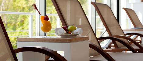 A sunlit room with a table set with a fruit bowl and a drink, overlooking a balcony with chairs.