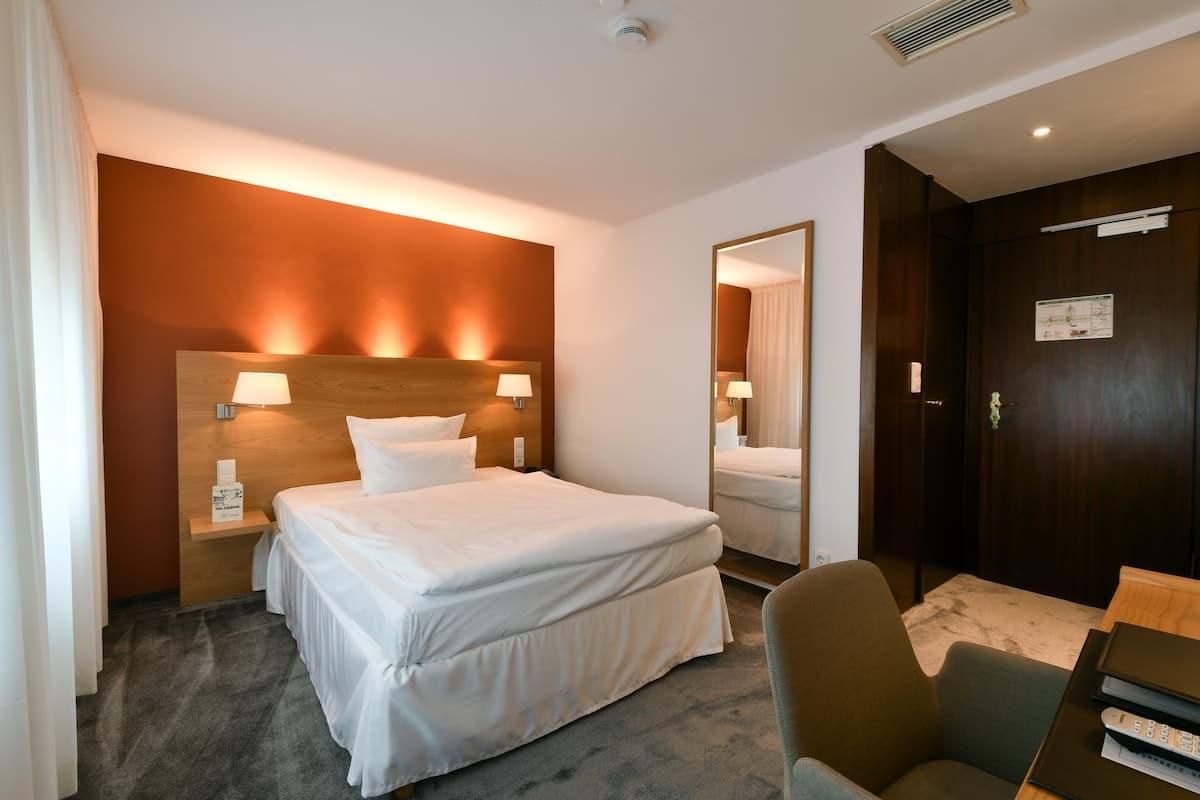 Quarto de hotel com cama grande, duas luminárias de cabeceira, cabeceira de madeira, espelho, escrivaninha com cadeira e porta com placa.
