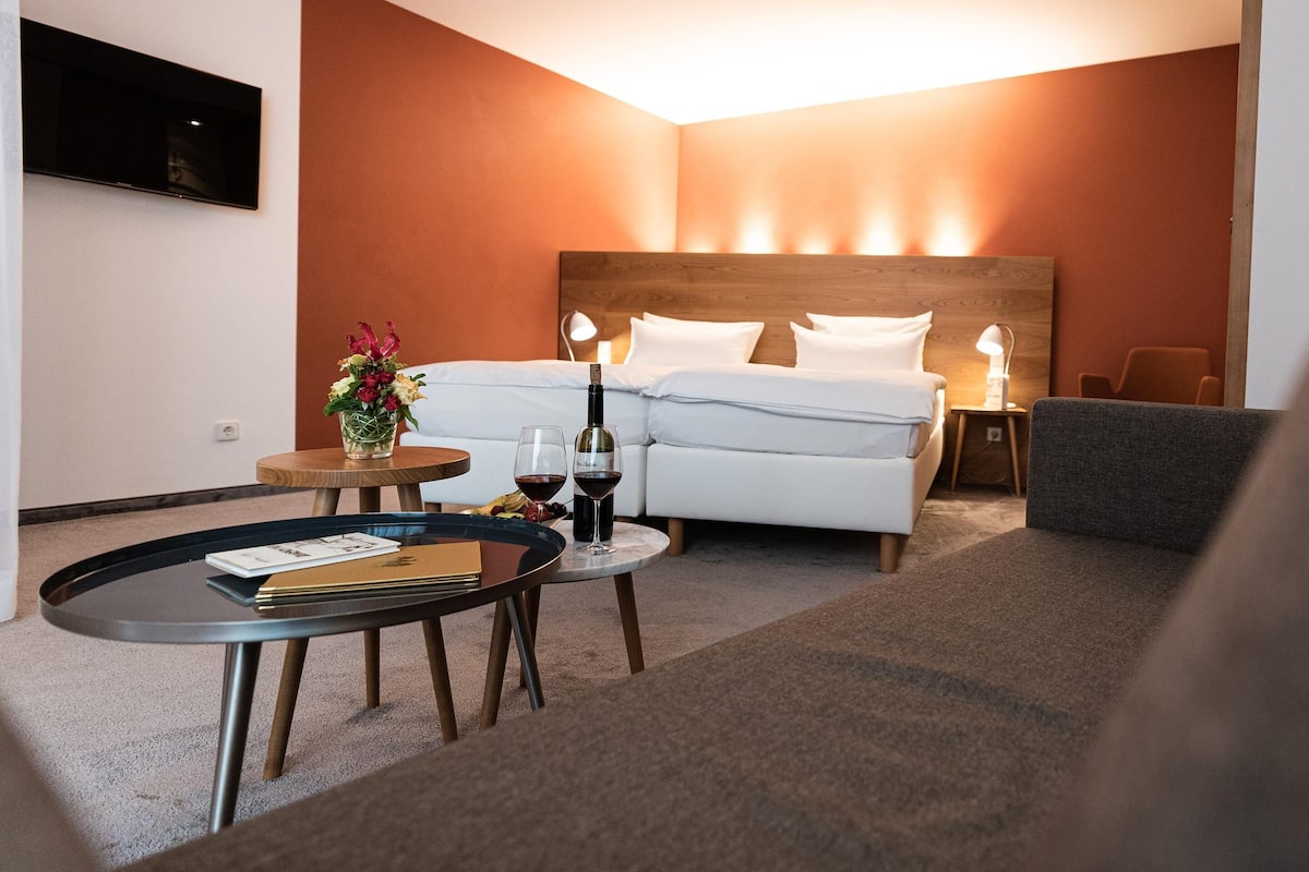Quarto de hotel com cama, mesa de cabeceira, luminária e uma mesinha redonda com uma garrafa de vinho e copos.