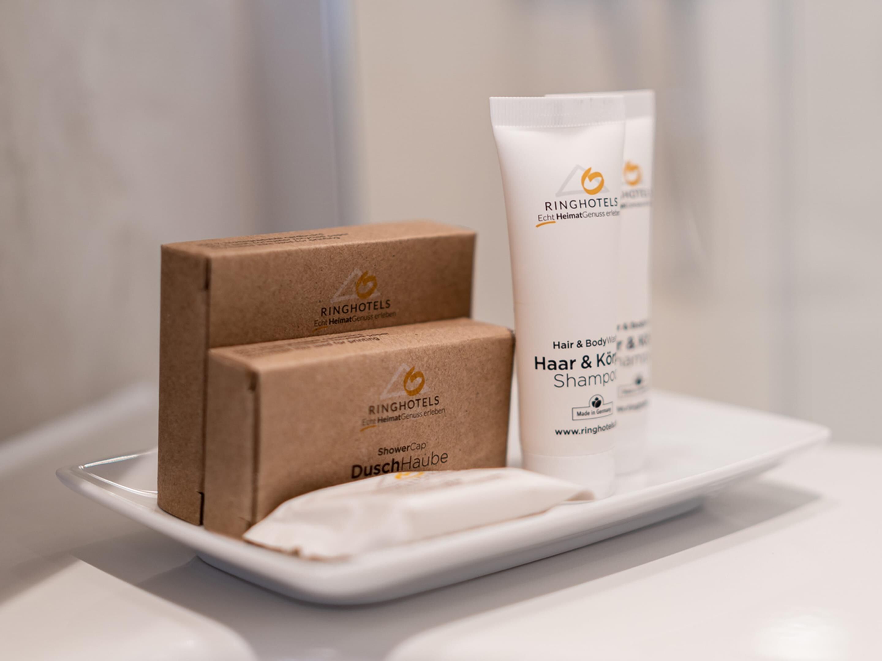Una encimera de baño con un plato blanco que sostiene un gorro de ducha, dos cajas marrones con la etiqueta 'RINGHOTELS' y un tubo blanco de 'Hair & Body Wash Shampoo'.