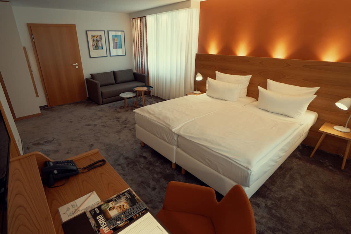 Quarto de hotel com cama, escrivaninha com telefone, cadeira, mesa pequena e abajur.