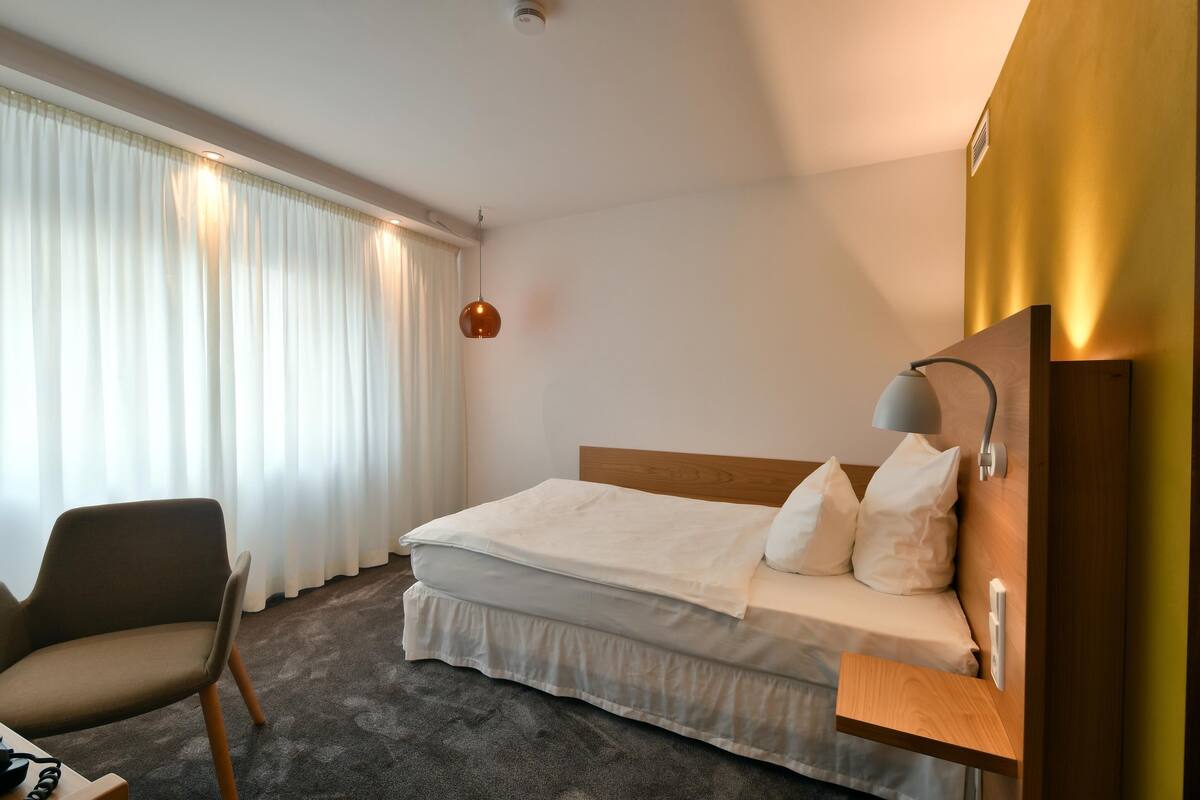 Quarto de hotel com cama, cadeira, mesa de cabeceira e abajur.