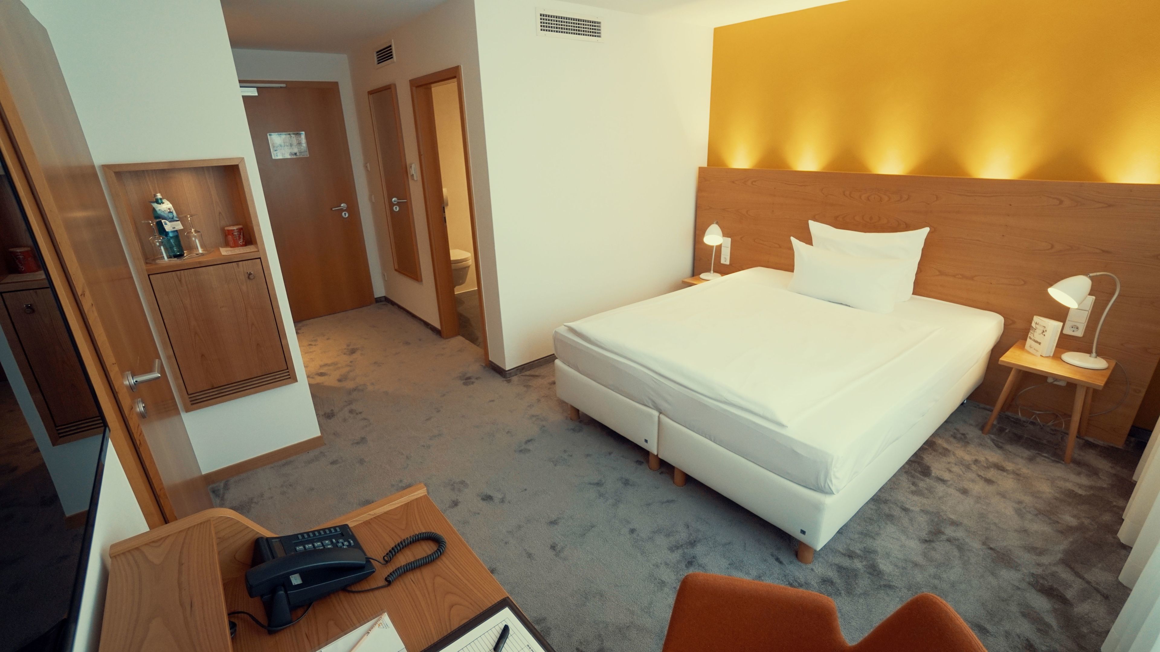 A hotel room with a bed, bedside lamps, a desk with a telephone, and a small cabinet.