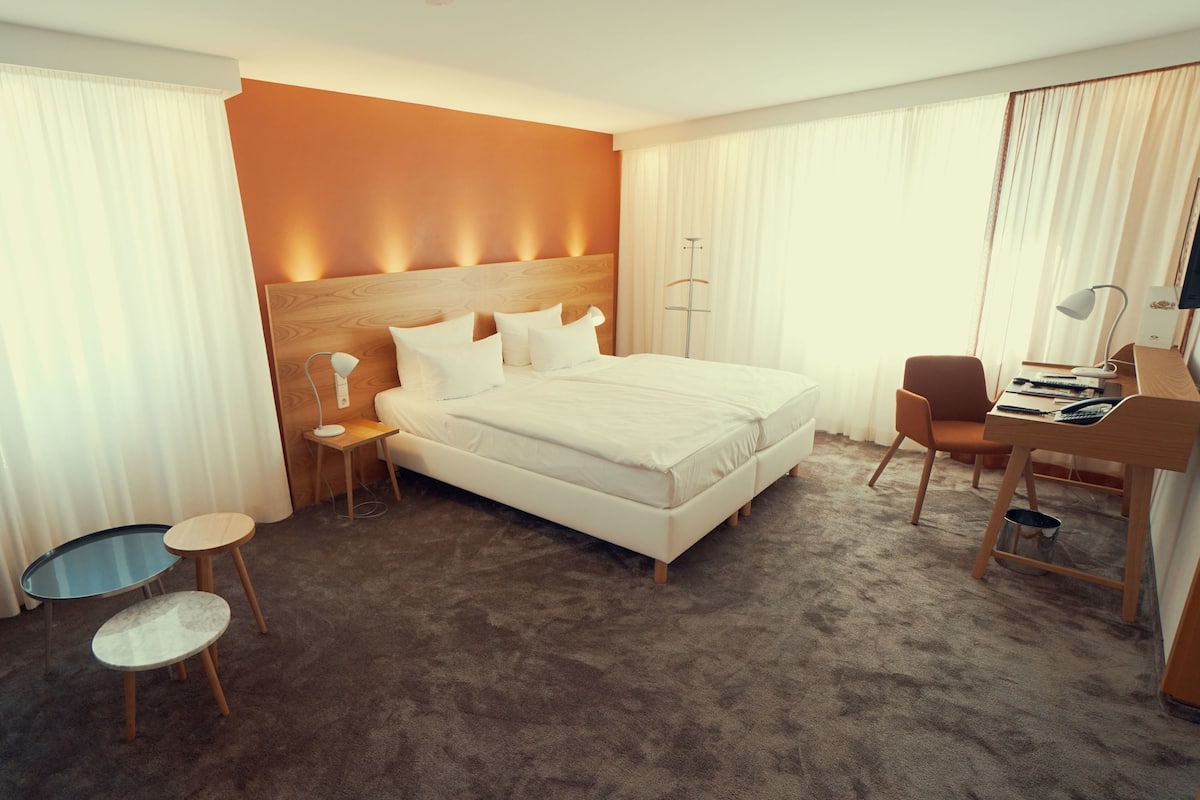 Quarto de hotel com uma cama grande, uma escrivaninha com cadeira, uma mesinha redonda e uma televisão.