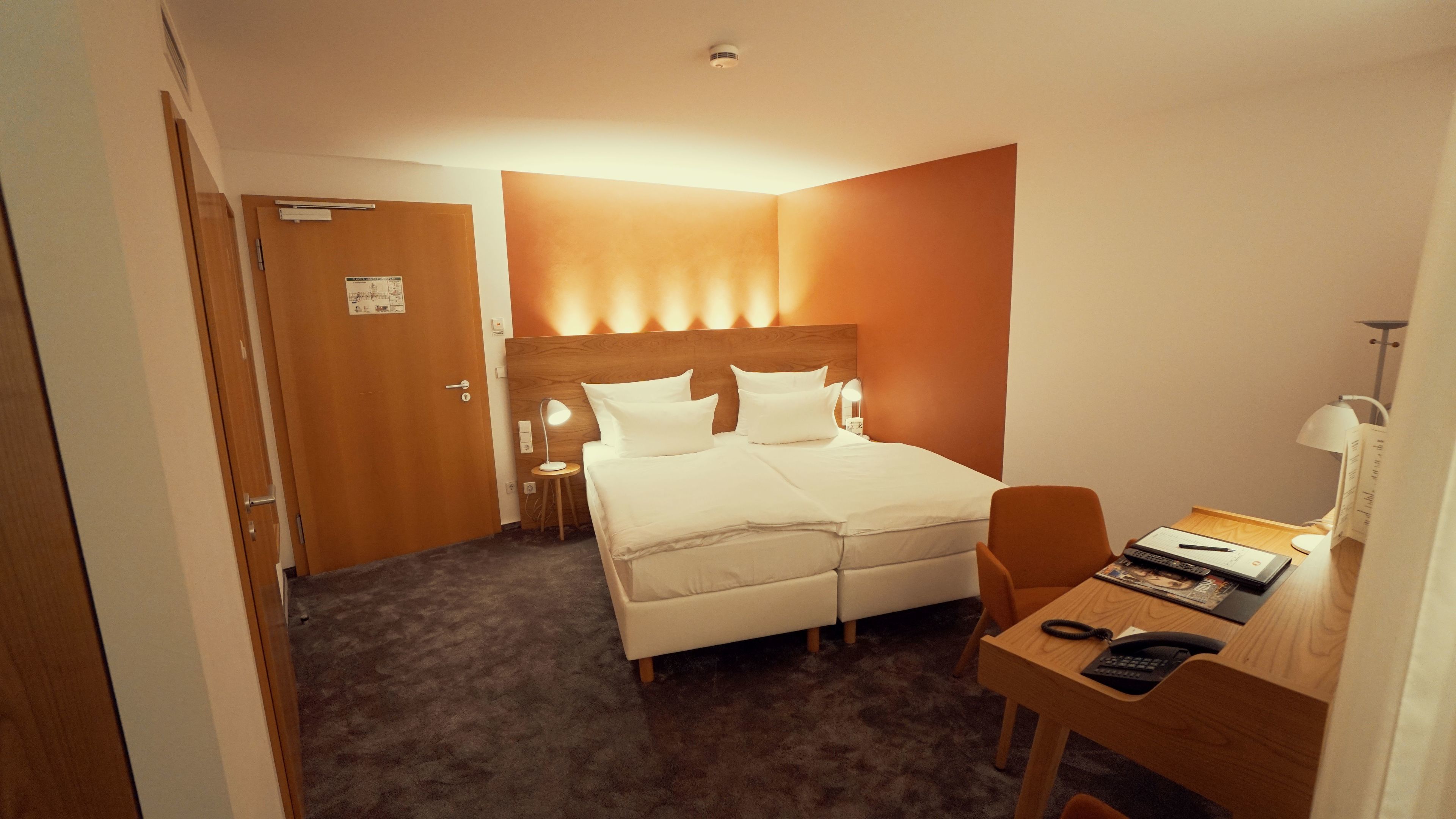 Una habitación de hotel con cama, mesita de noche, lámpara, escritorio, silla y una puerta.