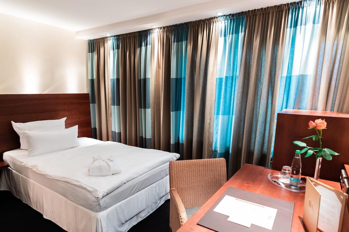 Quarto de hotel com uma cama, cabeceira de madeira, escrivaninha com um vaso de flores e janela com cortinas listradas.