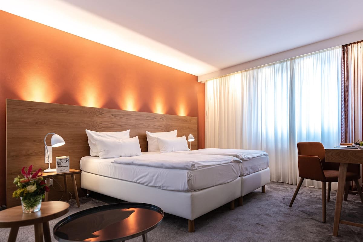 Quarto de hotel com uma cama grande, uma escrivaninha, uma cadeira, uma mesinha e uma mesa de cabeceira com abajur.